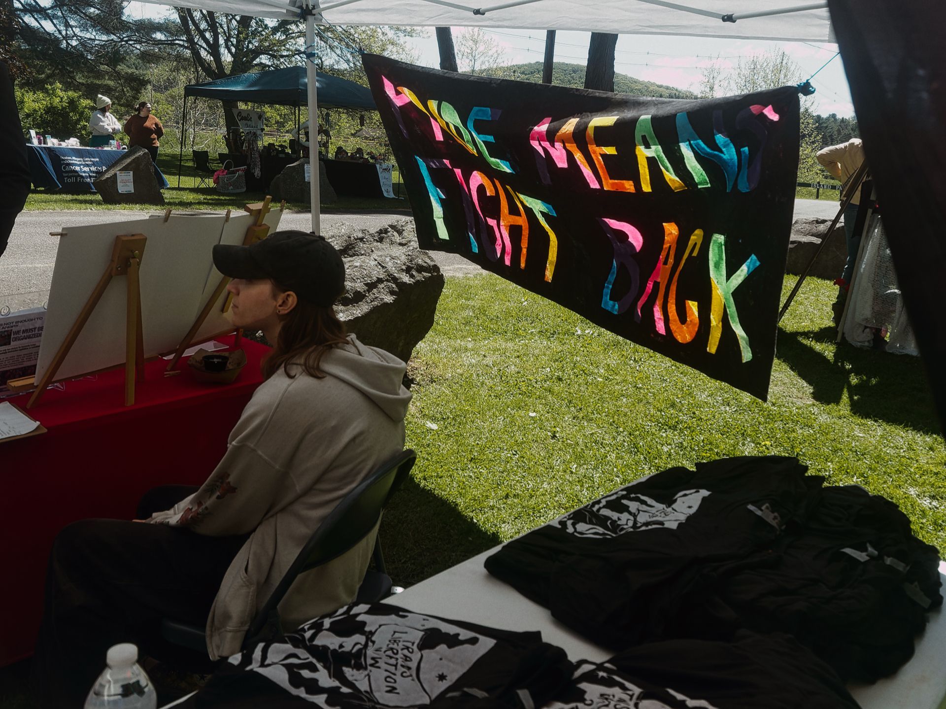 Outdoor event booth under a white canopy, dominated by a large black banner with multicolored hand-painted text reading "PRIDE MEANS FIGHT BACK." In the foreground, black t-shirts printed with "TRANS LIBERATION" are stacked on a table beside a water bottle. To the left, a person in a cream hoodie and black cap sits sideways at a red-draped table with easels behind them. A green lawn, trees, rolling hills, and a second teal canopy with other attendees are visible in the background.