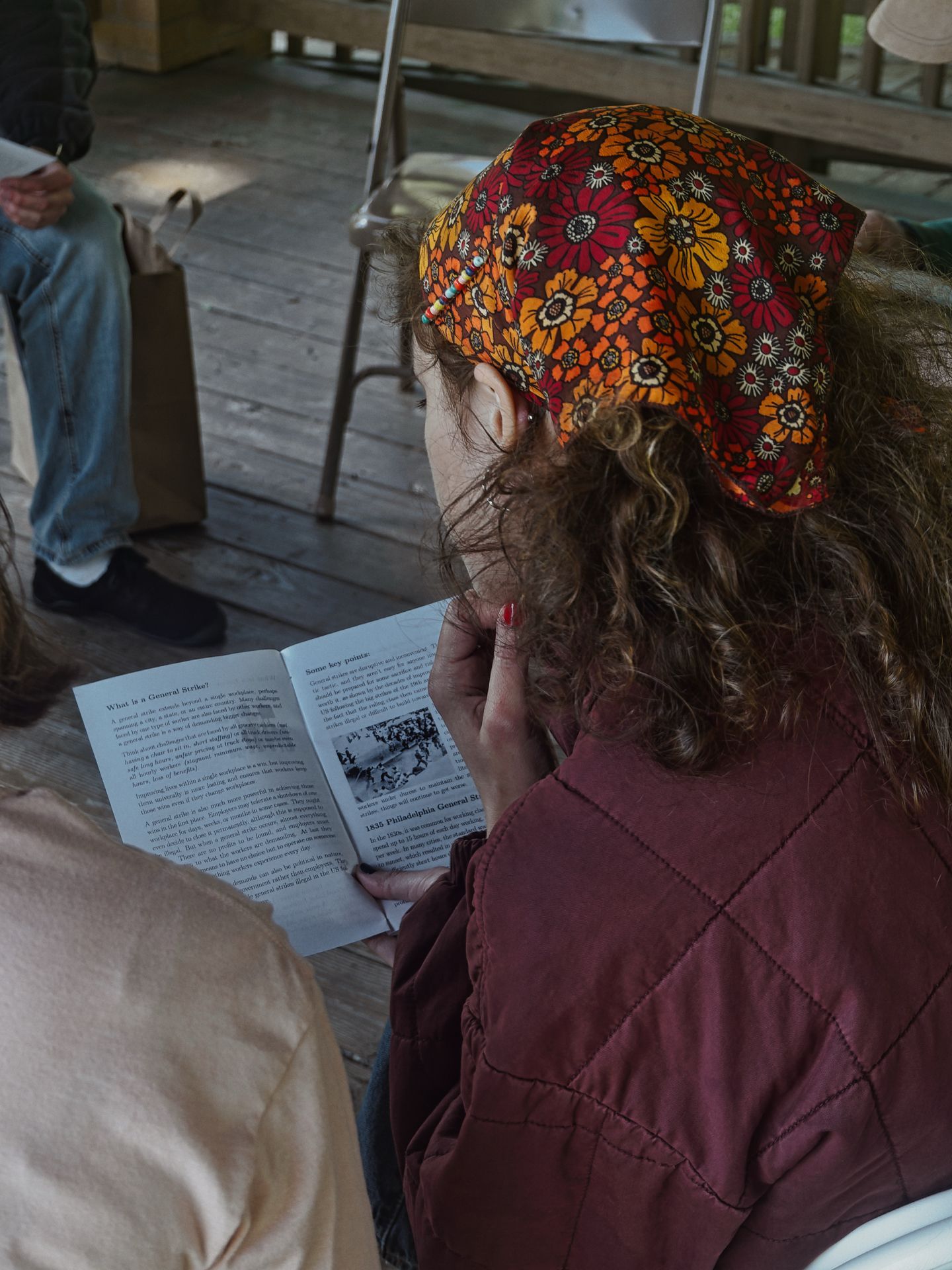 Woman with curly hair and a bold floral headscarf in orange, red, and gold reads a small pamphlet open to pages titled "What is a General Strike?" on the left and "Some key points:" on the right, with a black-and-white historical photograph visible on the right page. She wears a burgundy quilted jacket, her red-painted fingernail visible as she holds the booklet. A wooden deck setting with other seated figures blurred in the background.