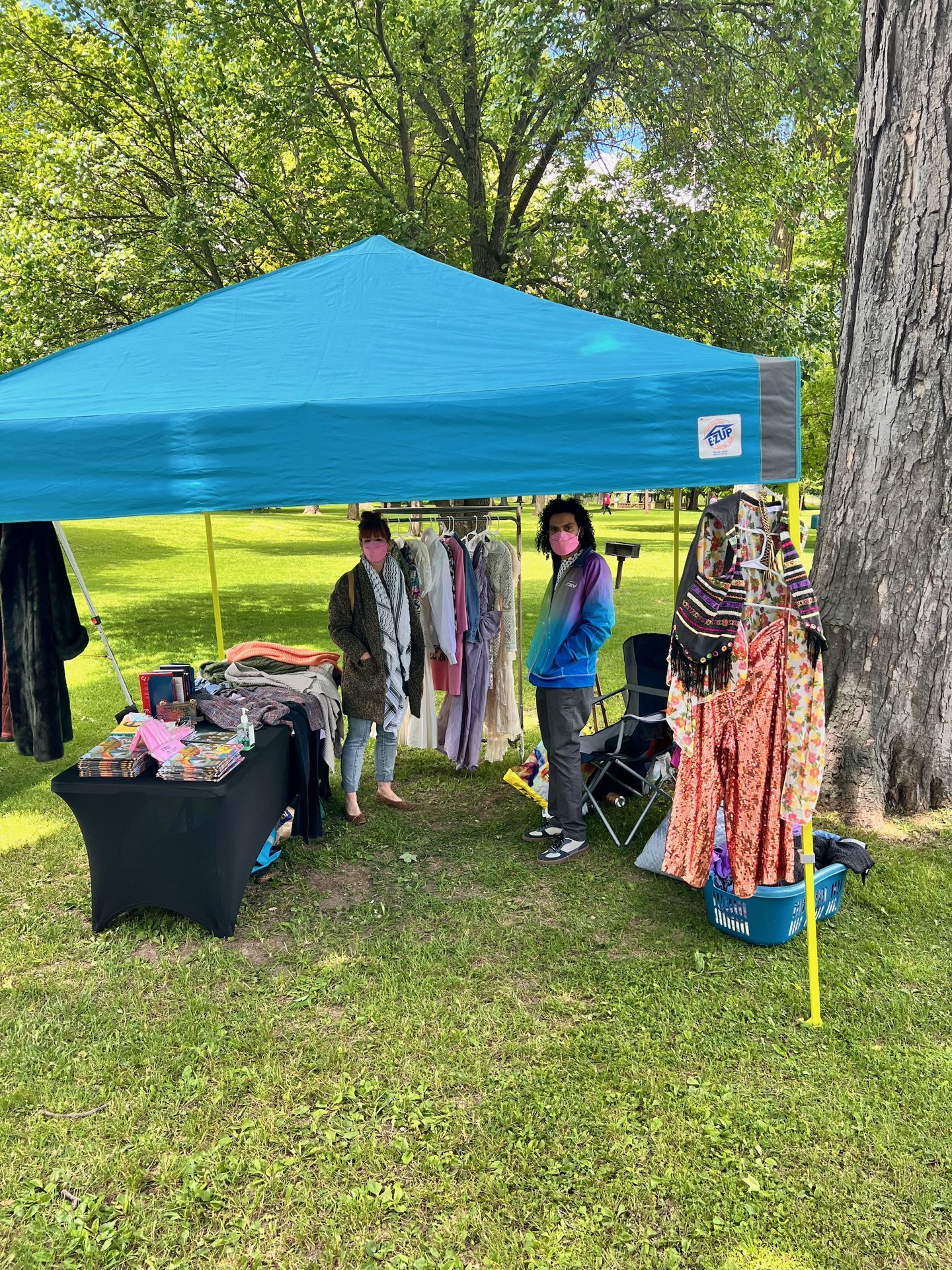 Two masked vendors stand beneath a teal pop-up canopy at an outdoor market in a grassy park, surrounded by hanging garments on racks — including flowing dresses and colorful patterned clothing — with a black-draped table of books and accessories to the left and a large tree trunk to the right.
