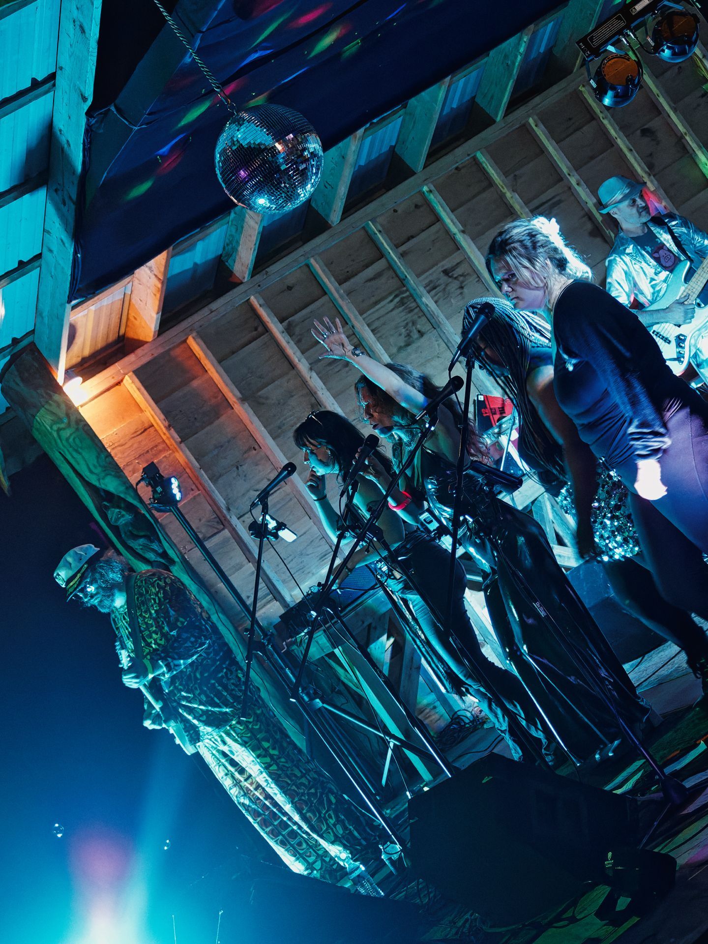 Vertically rotated shot of a band performing on a rustic wooden stage under teal-blue stage lighting, with a mirror disco ball hanging from the raftered ceiling above them — which reads as below due to the camera rotation. Several performers cluster around microphone stands at center, one with an arm raised, while a guitarist stands to the right and a figure in a patterned outfit occupies the far left. Stage monitors and equipment crowd the floor between them.
