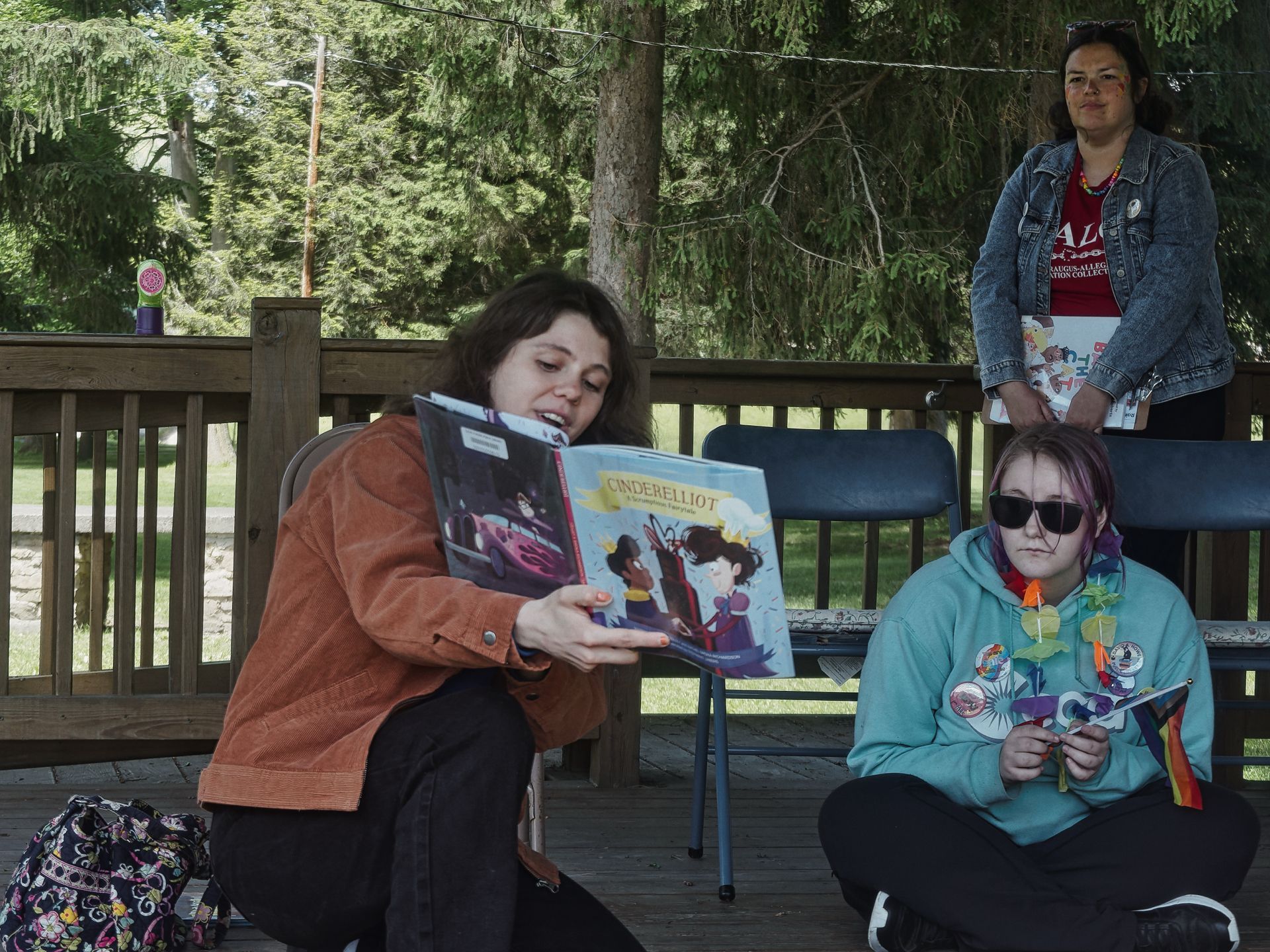On an outdoor wooden deck surrounded by trees, a woman in an orange corduroy jacket holds open a children's book titled *Cinderelliot: A Scrumptious Fairytale*, showing its illustrated cover to her audience. To her right, a young person in a teal hoodie sits cross-legged on the deck floor, wearing sunglasses and several enamel pins including a rainbow Pride flag pin, holding small items in their lap. In the background, a third person in a denim jacket and red shirt stands watching near the railing.