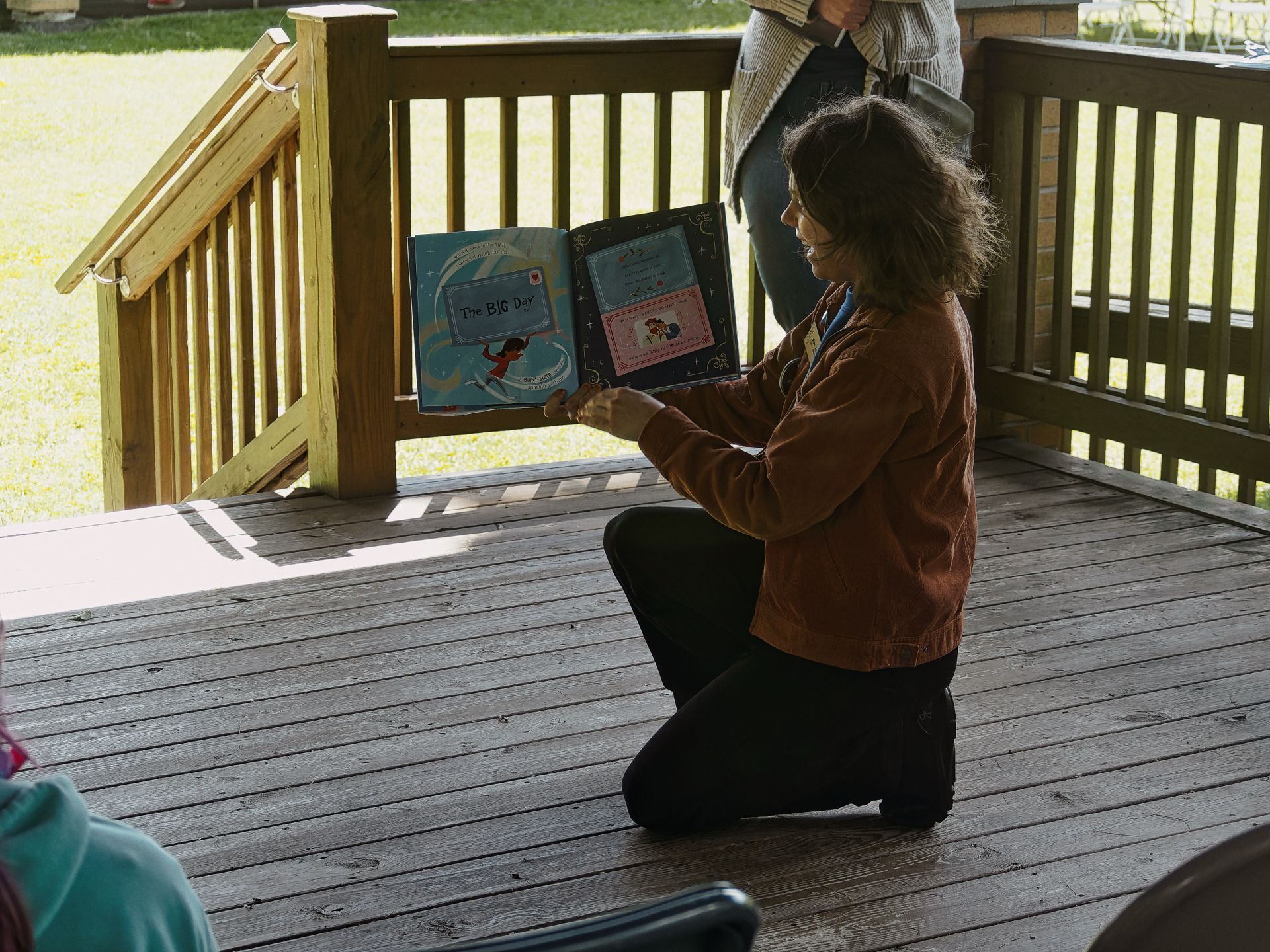 A woman in a rust-brown jacket kneels on a sunny wooden deck, holding open a children's picture book titled *The Big Day* to face an unseen audience. A second person stands behind her near the railing, partially cropped, with green grass visible through the wooden balusters in the background.