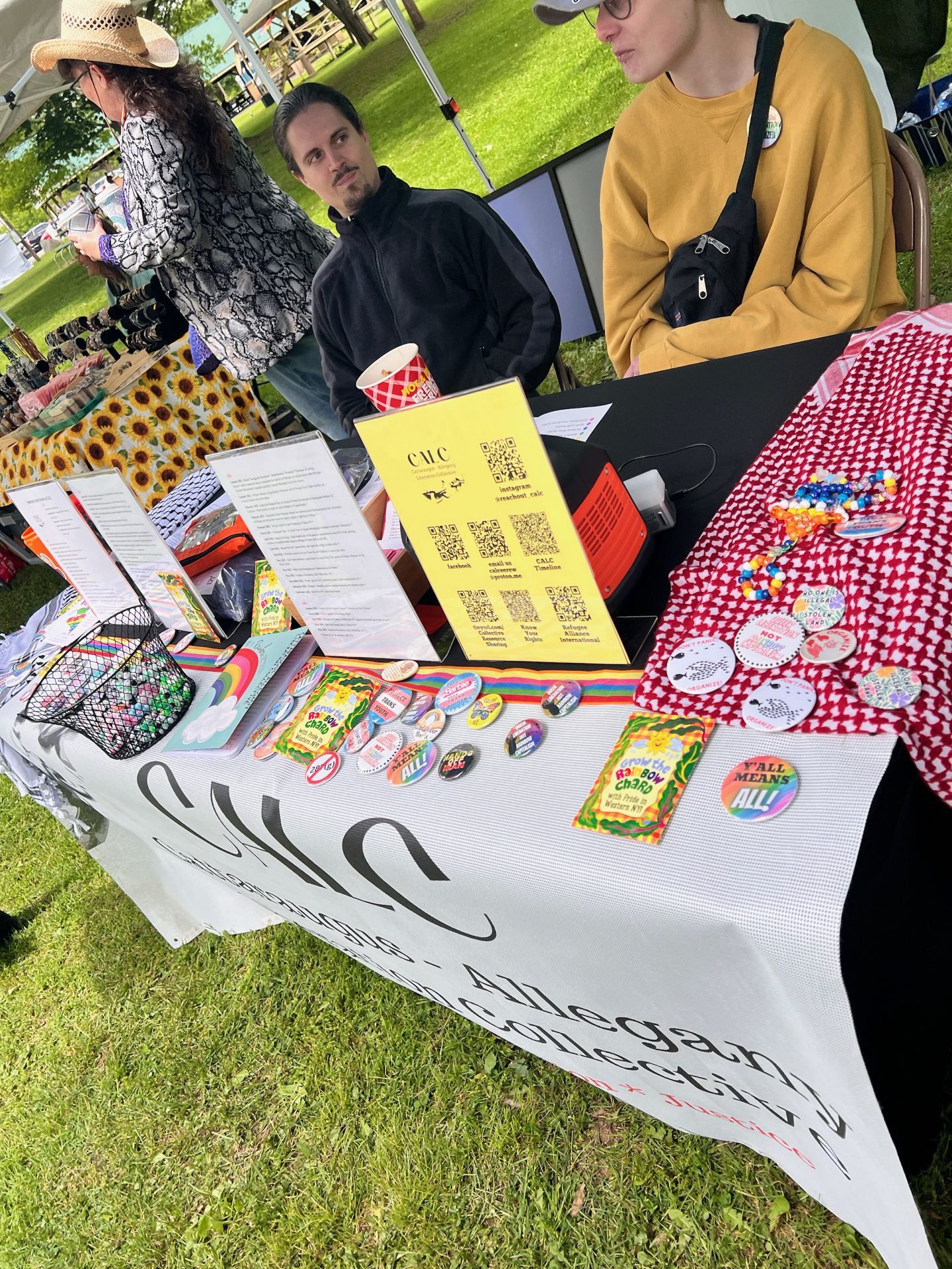 Outdoor festival booth for "Cattaraugus-Allegany Collective" — visible on a white tablecloth banner — covered in rainbow-themed pins and buttons, a yellow CALC sign with QR codes, scattered pamphlets, and a red-and-white checkered cloth on the right holding more buttons and beaded jewelry. Three people stand behind and beside the table: a woman in a wide-brim hat and snake-print top at left, a man in a black zip-up center, and a person in a mustard yellow sweatshirt at right, all set against a grassy park backdrop under a tent.