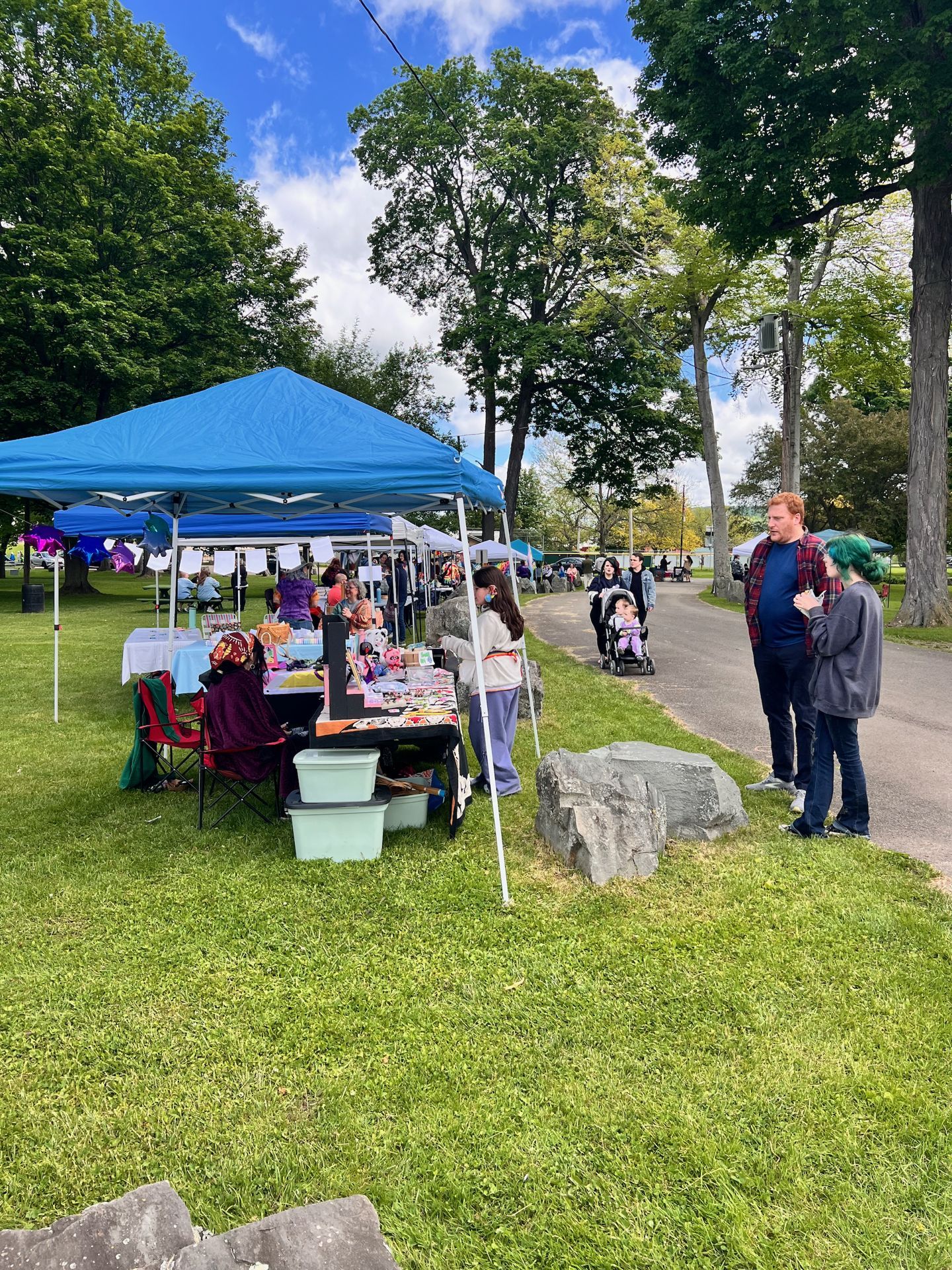 Vertical shot of an outdoor craft fair in a tree-lined park on a partly cloudy day, with a bright blue pop-up canopy shading a vendor table covered in colorful goods on the left, and two people — one with teal-dyed hair — conversing near large rocks at the edge of a paved path on the right, while a stroller and more visitors move through the background.