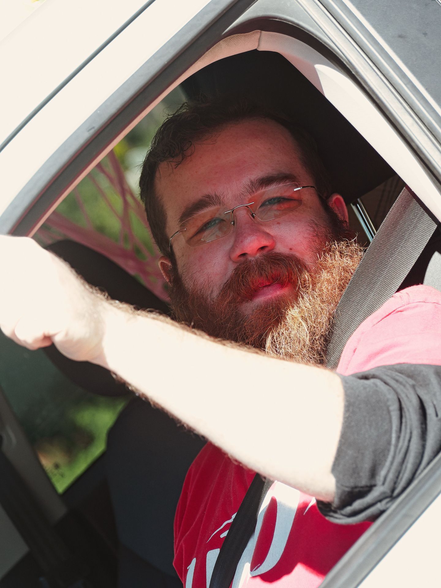 Bearded man with wire-rimmed glasses and a full auburn beard seated in the driver's seat of a vehicle, arm resting on the open window frame, seatbelt visible across his chest, wearing a red and grey layered shirt, with green foliage blurred softly in the background.