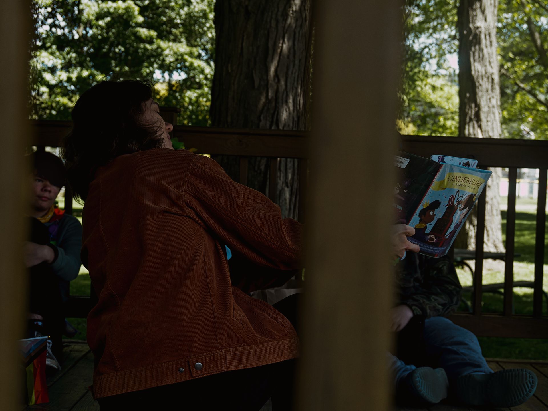 Shot from outside through the posts of a covered outdoor deck, a person in a rust-brown corduroy jacket turns their head upward while a child to their right holds up a copy of *Cinderella* — its illustrated cover showing a dark-skinned figure and a rabbit visible in the dim light. A second person in a yellow scarf sits to the left, with sunlit trees and a grassy park blurred in the background.
