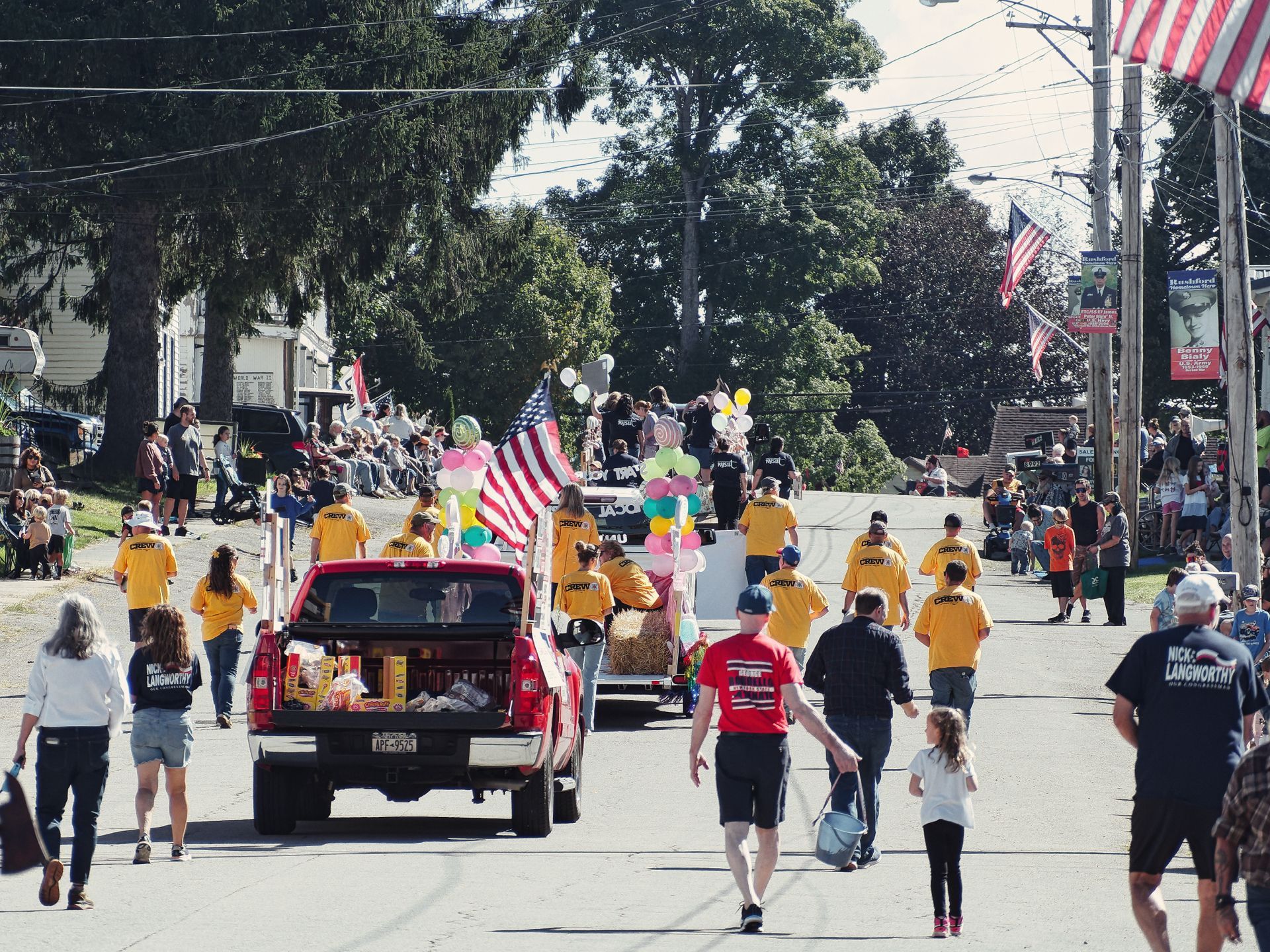 Small-town parade moving down a tree-lined street, with a red pickup truck towing a float decorated with balloons, hay bales, and an American flag, surrounded by volunteers in matching yellow "CREW" shirts. Spectators line the sidewalks on both sides, a "NICK LANGWORTHY" campaign shirt is visible on the right foreground, and a utility pole to the right displays an American flag and a political banner reading "Benny Blaky."