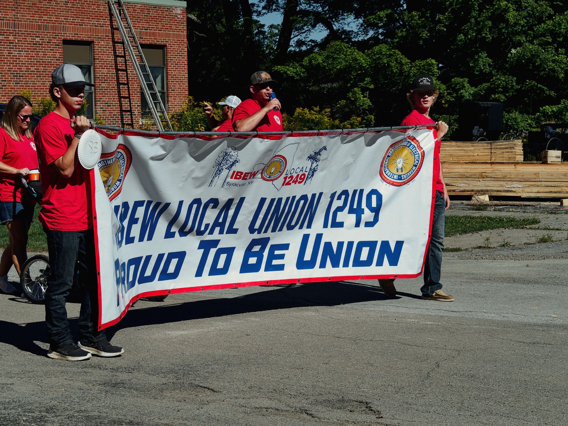 Group of people in matching red t-shirts marching in a parade, carrying a large white banner with red trim reading "IBEW LOCAL UNION 1249 / PROUD TO BE UNION" with union logos and "Syracuse, NY" visible at the top. A man at center-rear speaks into a microphone, while a brick building with a ladder and dense green trees fill the background.