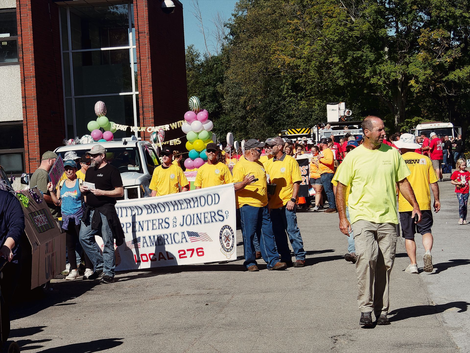 Outdoor labor day parade along a street, with a group of people in yellow shirts carrying a white banner reading "United Brotherhood of Carpenters and Joiners of America — Local 276," an American flag printed at center. Behind them, a decorated float with colorful balloons and a sign reading "Start Your Career With [Carpenters]" follows the procession, with a red brick building to the left and dense trees lining the background under a clear blue sky.