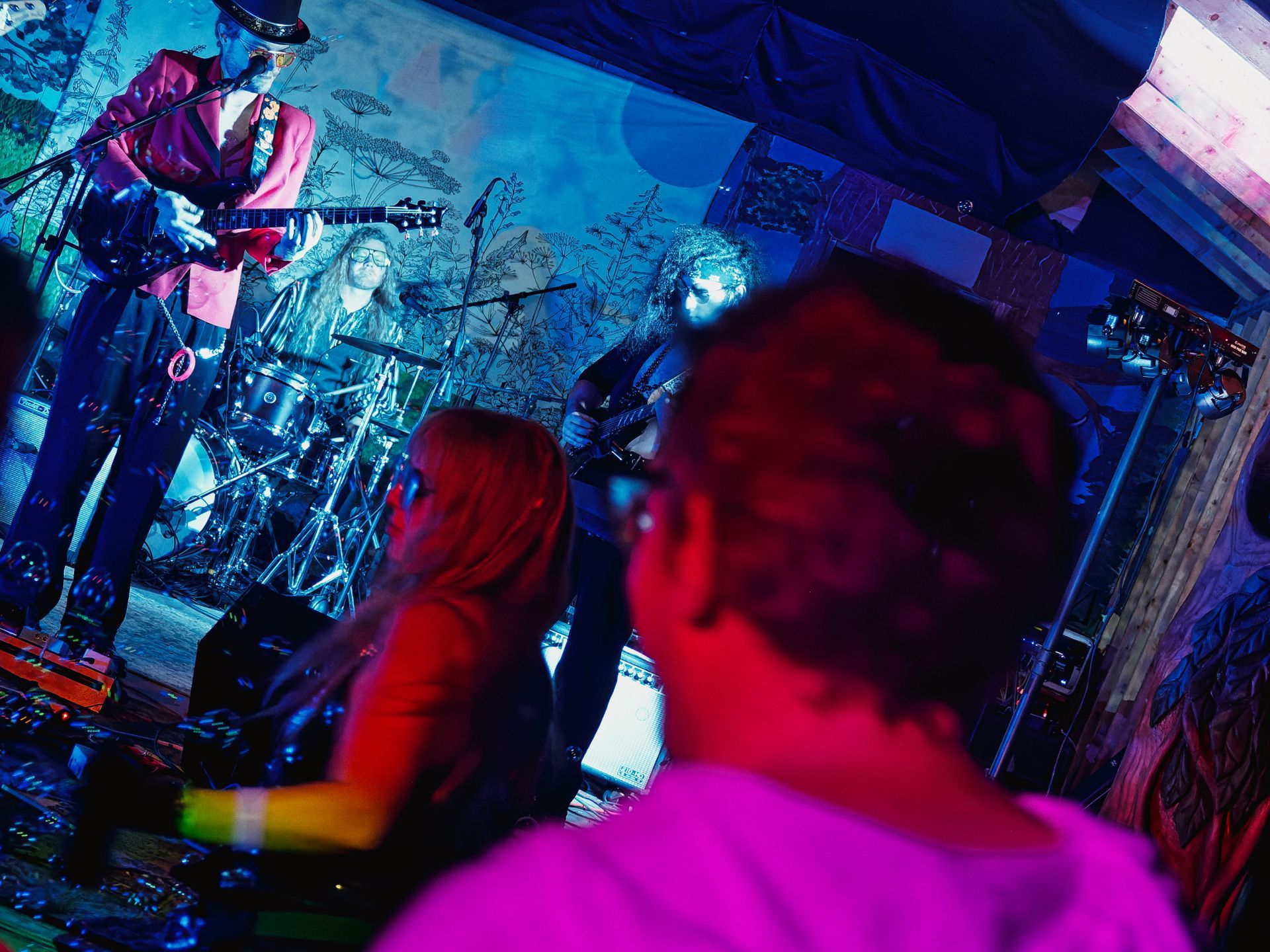 Two audience members — one in magenta, one in red — watch a three-piece band performing on a small, intimately lit stage, the foreground figures blurred in shallow depth. The guitarist on the left wears a pink blazer and top hat, a drummer sits center behind a full kit, and a bassist stands to the right, all bathed in deep blue stage light against a painted botanical backdrop.