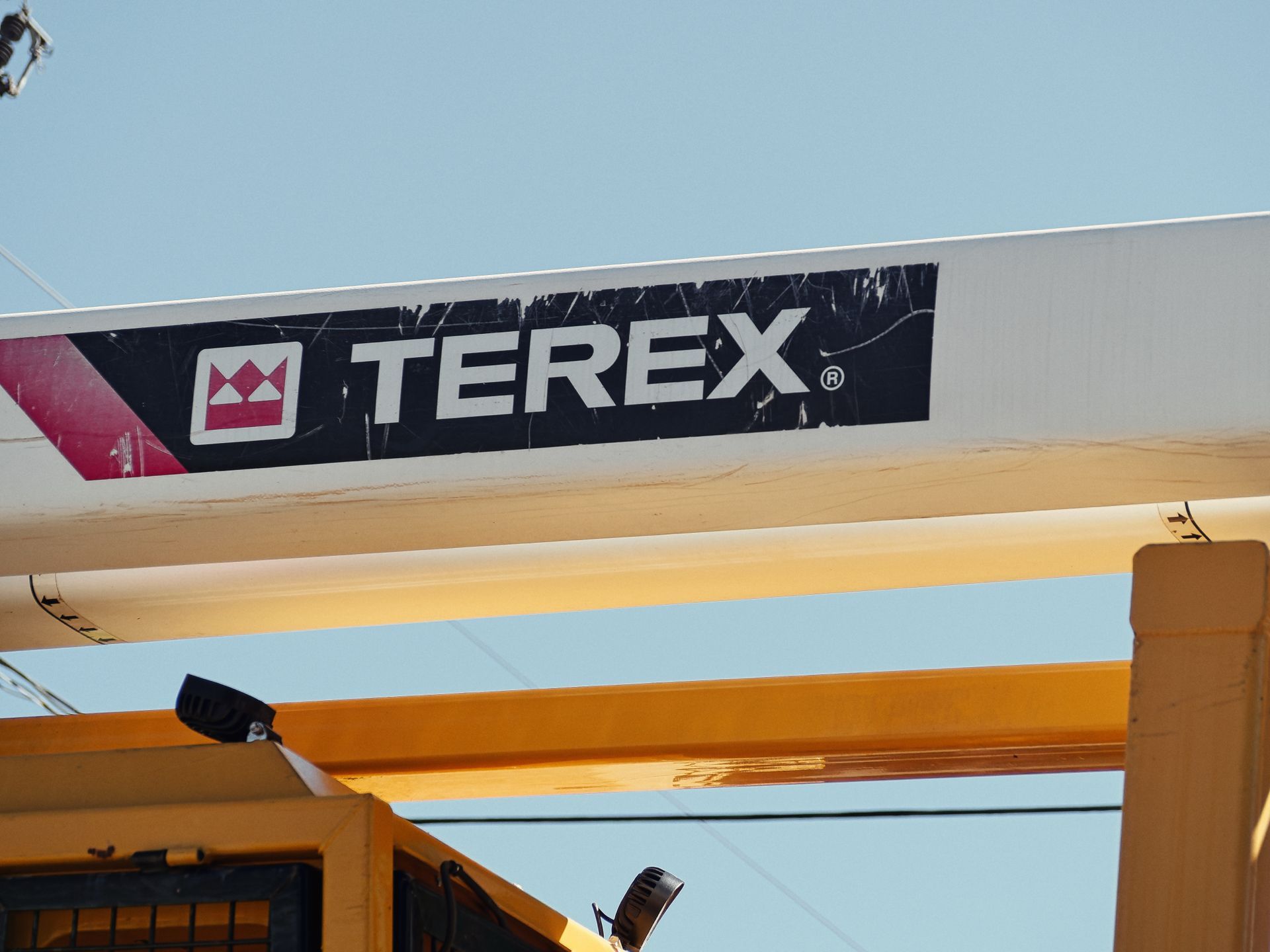 A weathered "TEREX®" logo decal — white text on a scuffed dark navy rectangle with a red crown emblem — affixed to a white beam of yellow heavy construction equipment, shot from below against a pale blue sky.