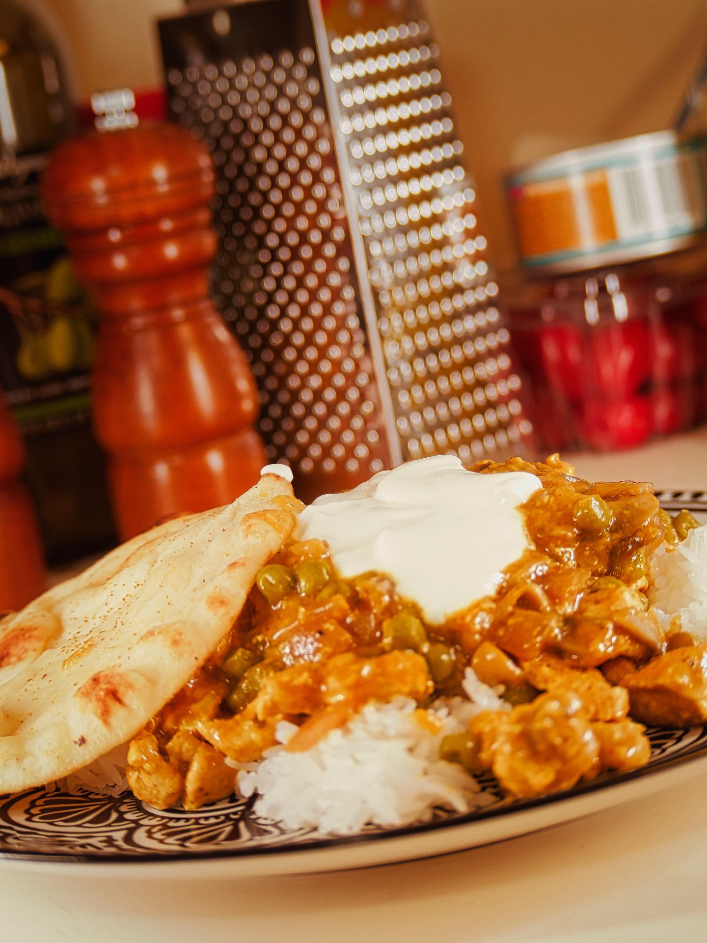 Vertical shot of a patterned ceramic plate piled with white rice, an orange-brown curry with peas and chunks of chicken, a dollop of white yogurt or sour cream, and a flatbread naan leaning to the left. A wood pepper mill, box grater, and glass jars sit blurred in the shallow depth of the background.