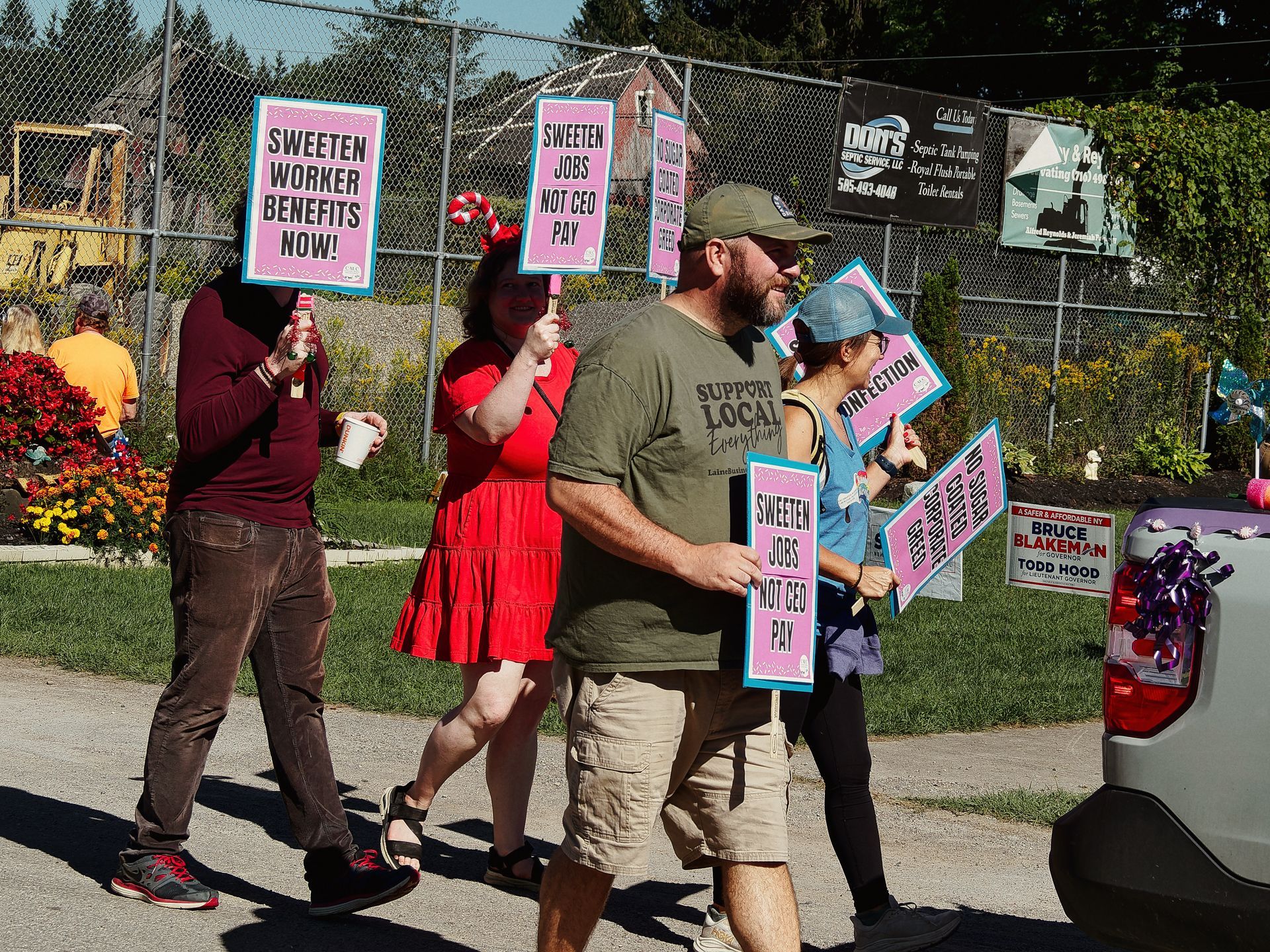 A small group of protesters marching along a street, holding pink and teal picket signs reading "SWEETEN WORKER BENEFITS NOW!" and "SWEETEN JOBS NOT CEO PAY." The man in the foreground wears an olive t-shirt reading "Support Local Everything" and khaki shorts; behind him, a woman in a red dress holds a candy cane-striped sign handle. A white vehicle to the right bears a "Bruce Blakeman / Todd Hood" campaign sign, and a "Don's Septic Service" billboard is visible on the chain-link fence in the background.