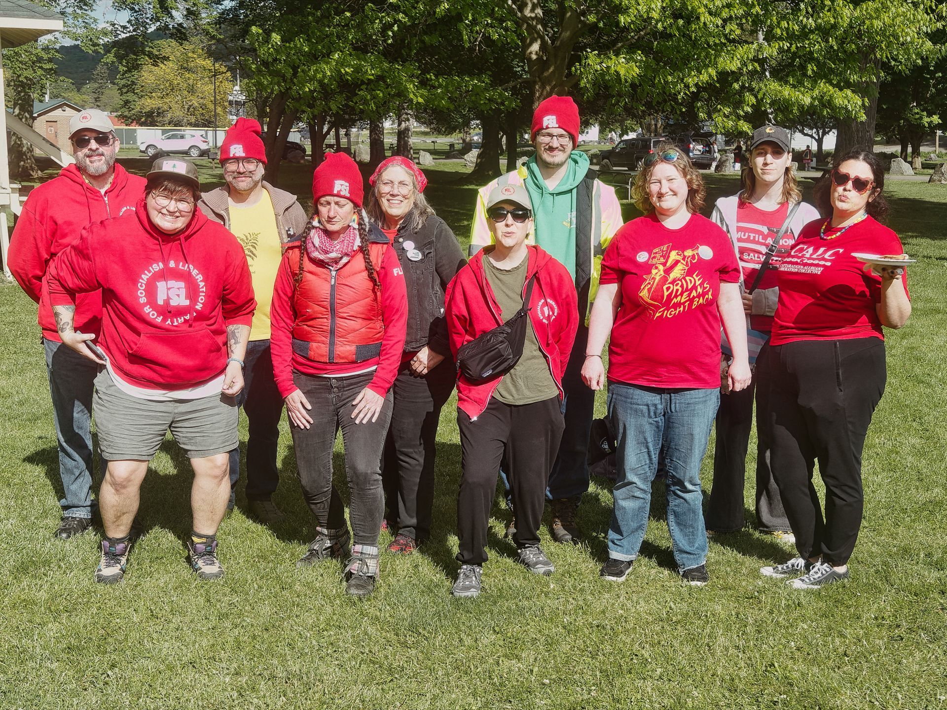 Group of about ten people posing together on a sunny grass lawn in a park, most wearing red — hoodies, jackets, and beanies bearing "PSL" logos. The leftmost person's red hoodie reads "PARTY FOR SOCIALISM & LIBERATION," and a red shirt in the center-right reads "PRIDE MEANS FIGHT BACK." Trees and parked cars blur softly in the background.