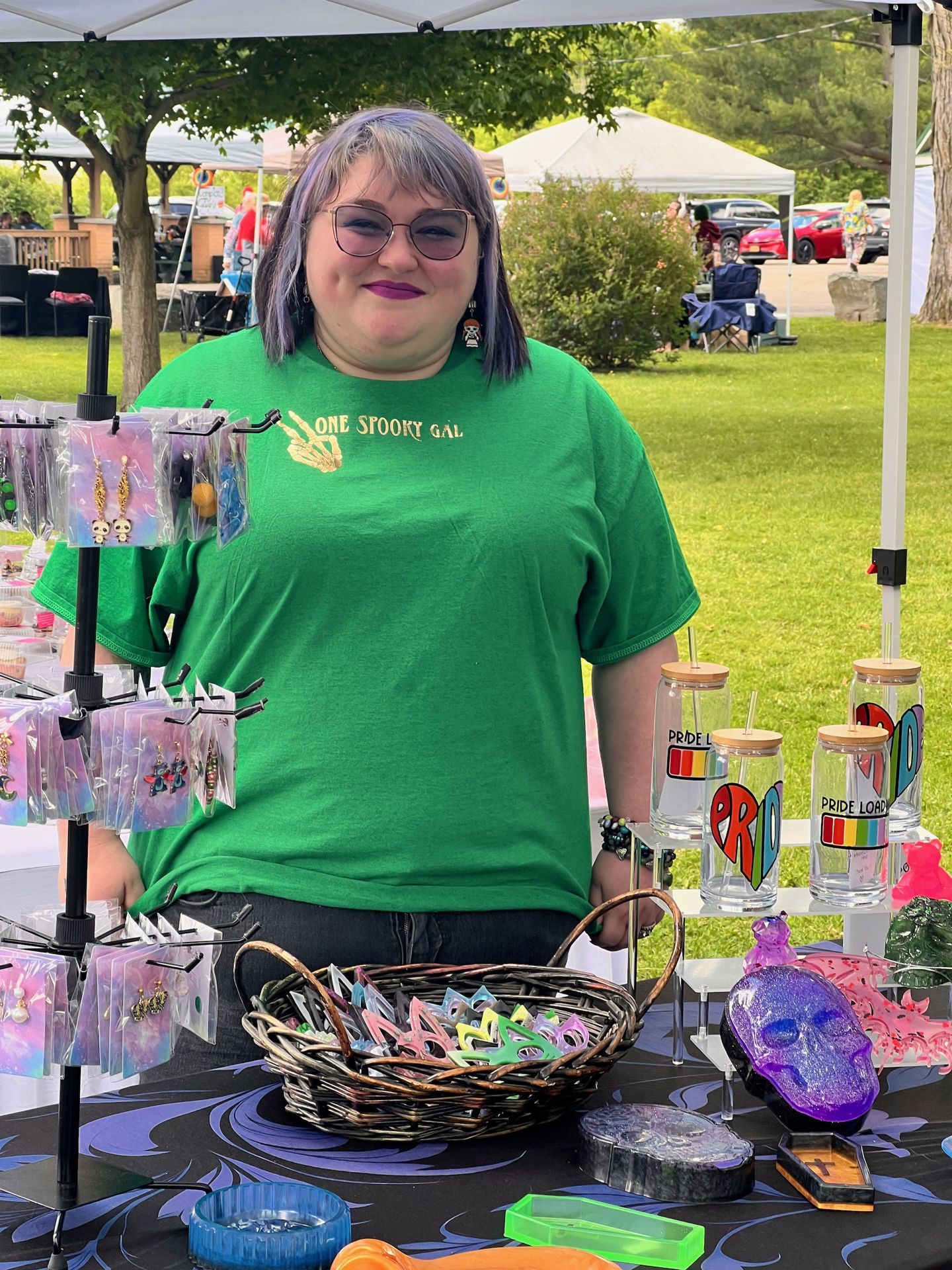 Vendor with lavender hair and glasses wearing a green t-shirt reading "ONE SPOOKY GAL" stands behind a craft fair table covered in handmade jewelry on display racks, a wicker basket of colorful hair clips, glass can tumblers labeled "PRIDE LOAD" with rainbow designs, and a purple resin skull — a white canopy tent and green park lawn visible in the background.