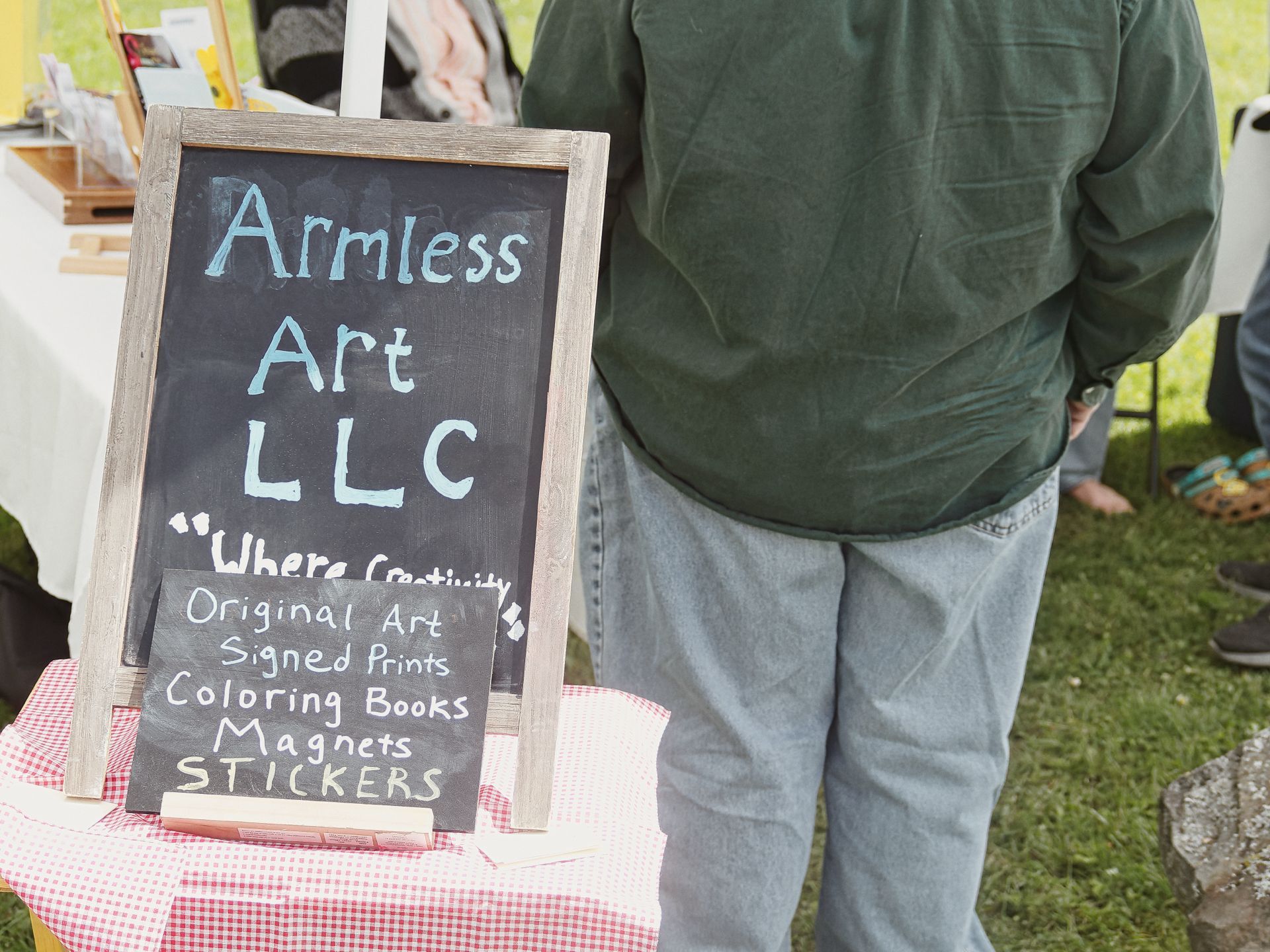 Chalkboard sign reading "Armless Art LLC — 'Where Creativity' / Original Art / Signed Prints / Coloring Books / Magnets / STICKERS" propped on a red-and-white gingham tablecloth, with a person in a dark green jacket and jeans standing with their back to the camera just to the right, at what appears to be an outdoor craft fair.