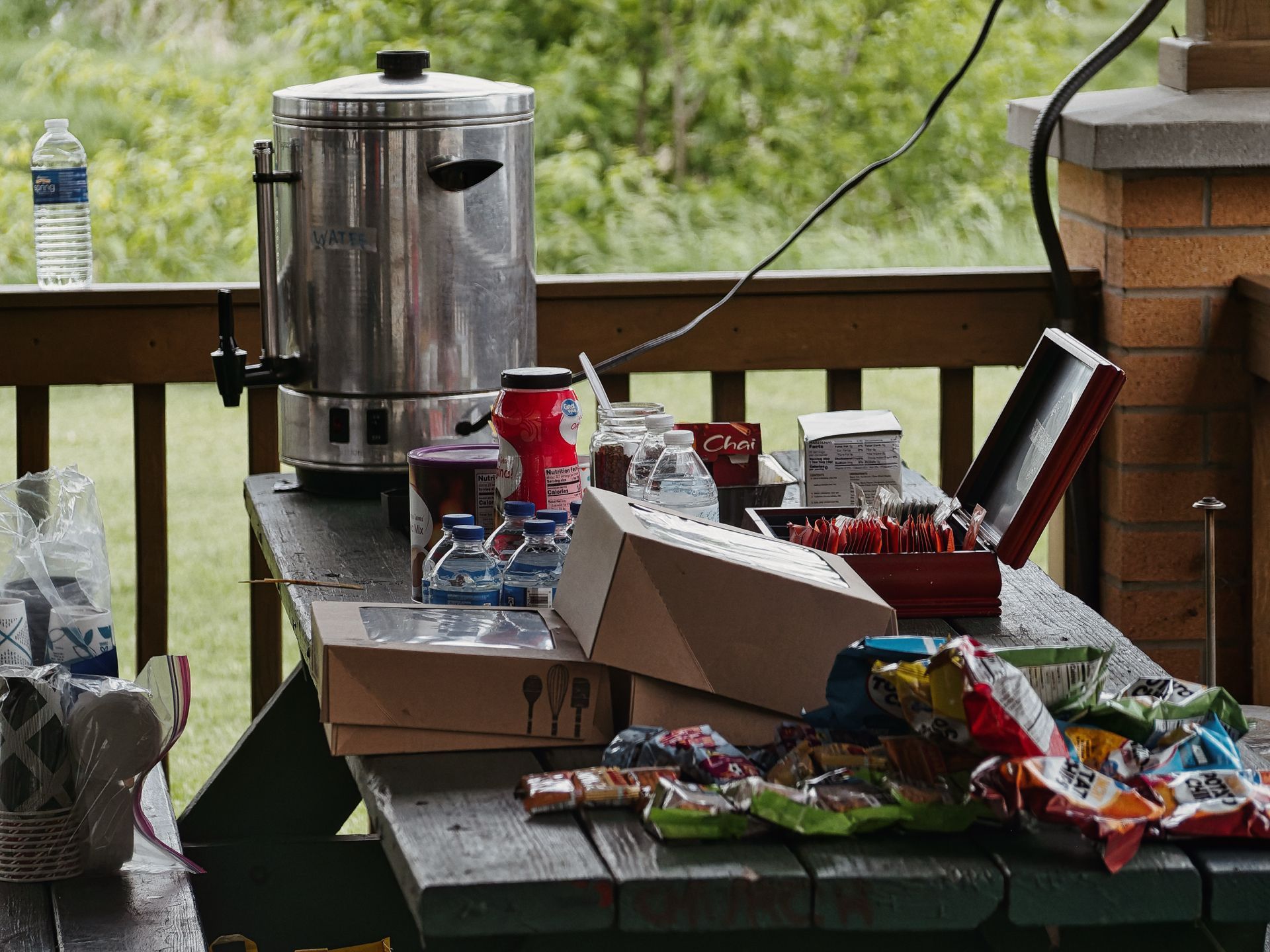 A cluttered outdoor deck table loaded with a large stainless steel hot water urn labeled "WATER," a red canister, mini water bottles, a box of Chai tea packets, an open cardboard box, a red tin filled with sugar packets, and an assortment of chip bags and snack wrappers spread across the foreground. A wicker basket with utensils sits to the left, and a lone water bottle stands on the railing behind the urn, with green trees blurred softly in the background.