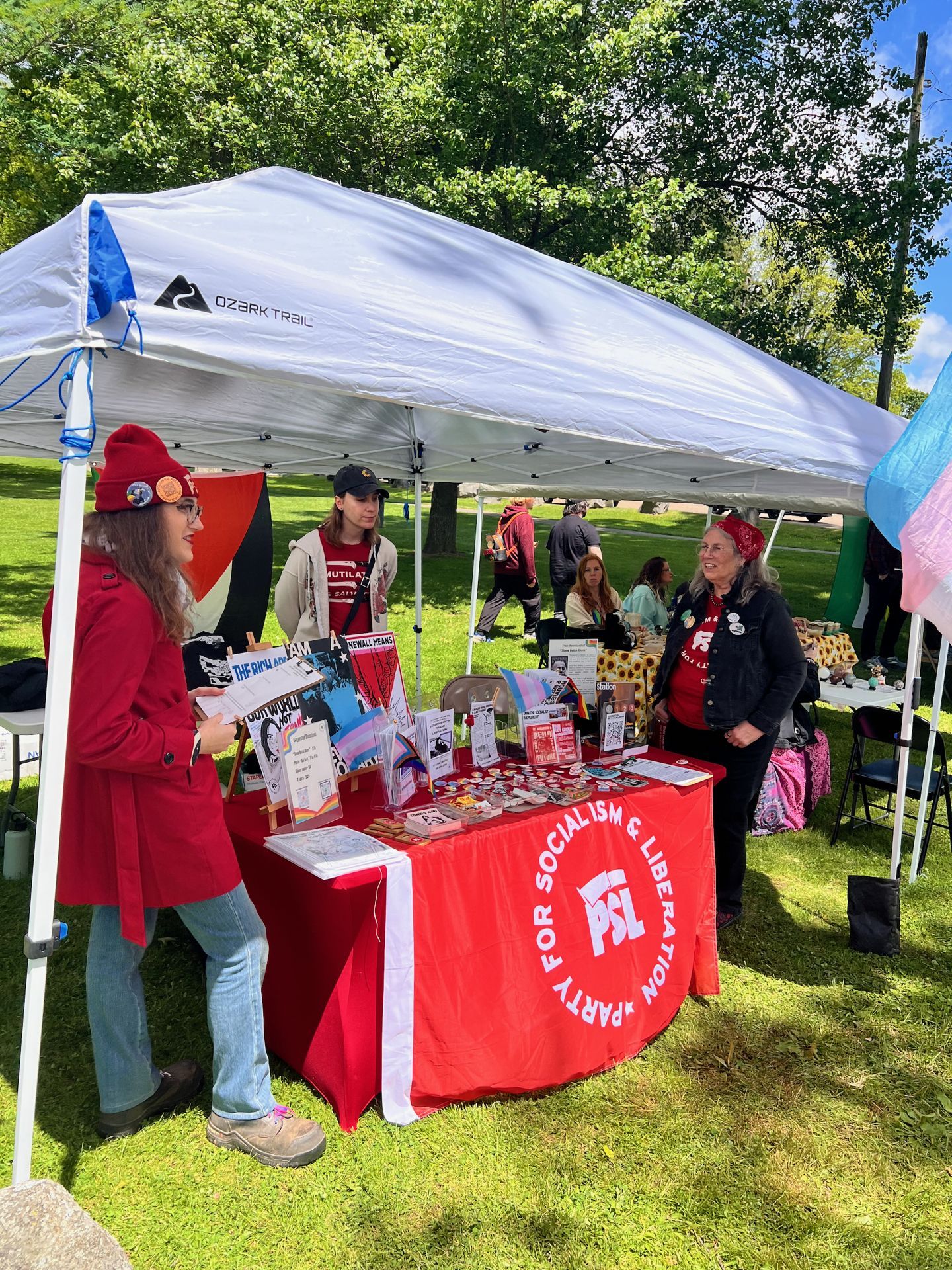 Vertical shot of an outdoor tabling event under a white Ozark Trail canopy on a grassy park lawn, with a red tablecloth reading "PARTY FOR SOCIALISM & LIBERATION" and the PSL logo across the front, covered in pins, pamphlets, and rainbow-themed materials. On the left, a person in a red coat and red beanie holds a sign; across the table, an older person in a red shirt and red bandana smiles; a third person in a black cap stands between them. A trans pride flag is partially visible at the far right edge, and a Palestinian flag hangs at the back of the tent.