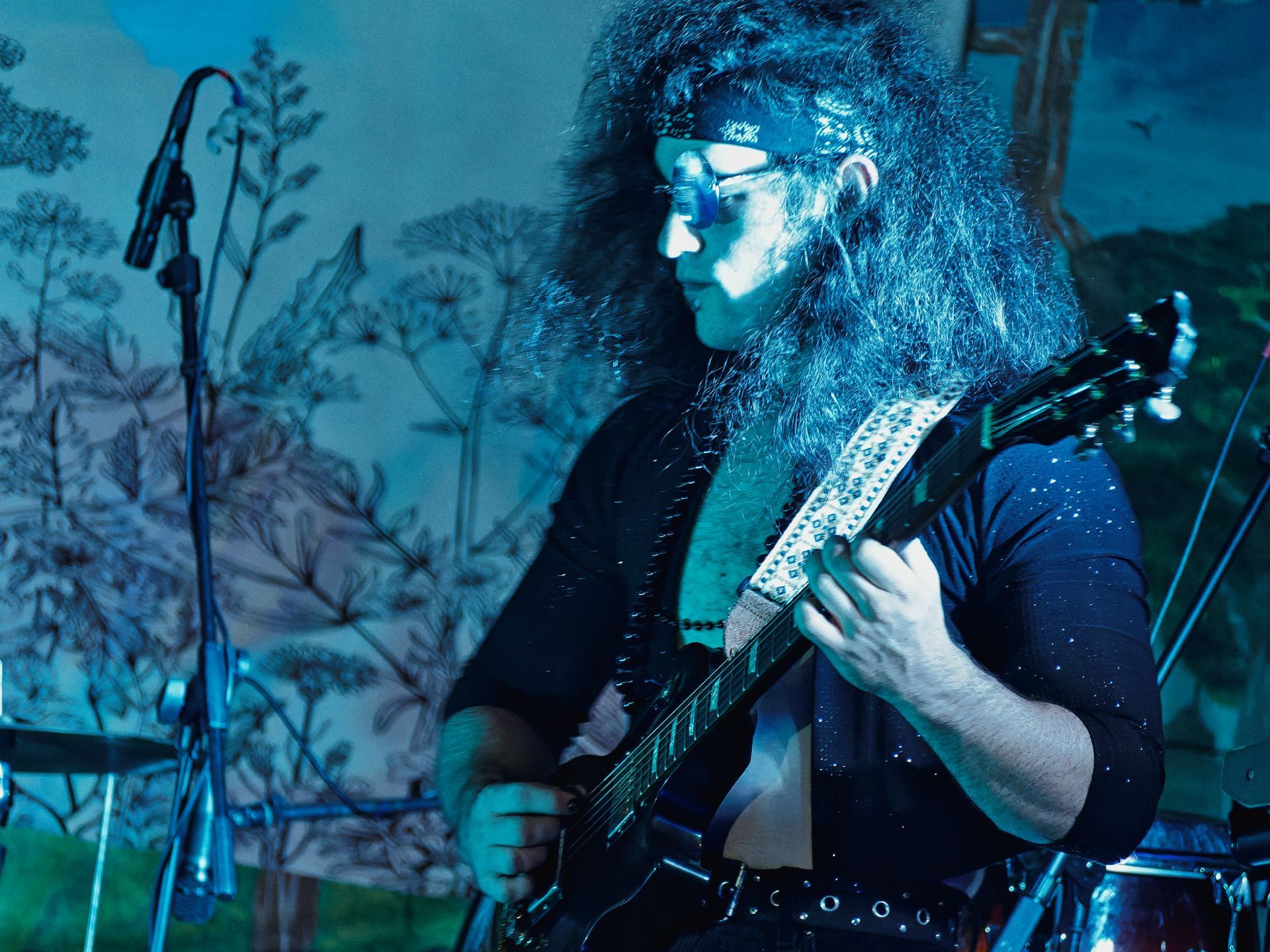Long-haired guitarist wearing round glasses and a bandana, bathed in deep blue stage lighting, playing a dark electric guitar with a studded strap. A microphone stand rises to the left, and a painted mural of trees and sky fills the background.