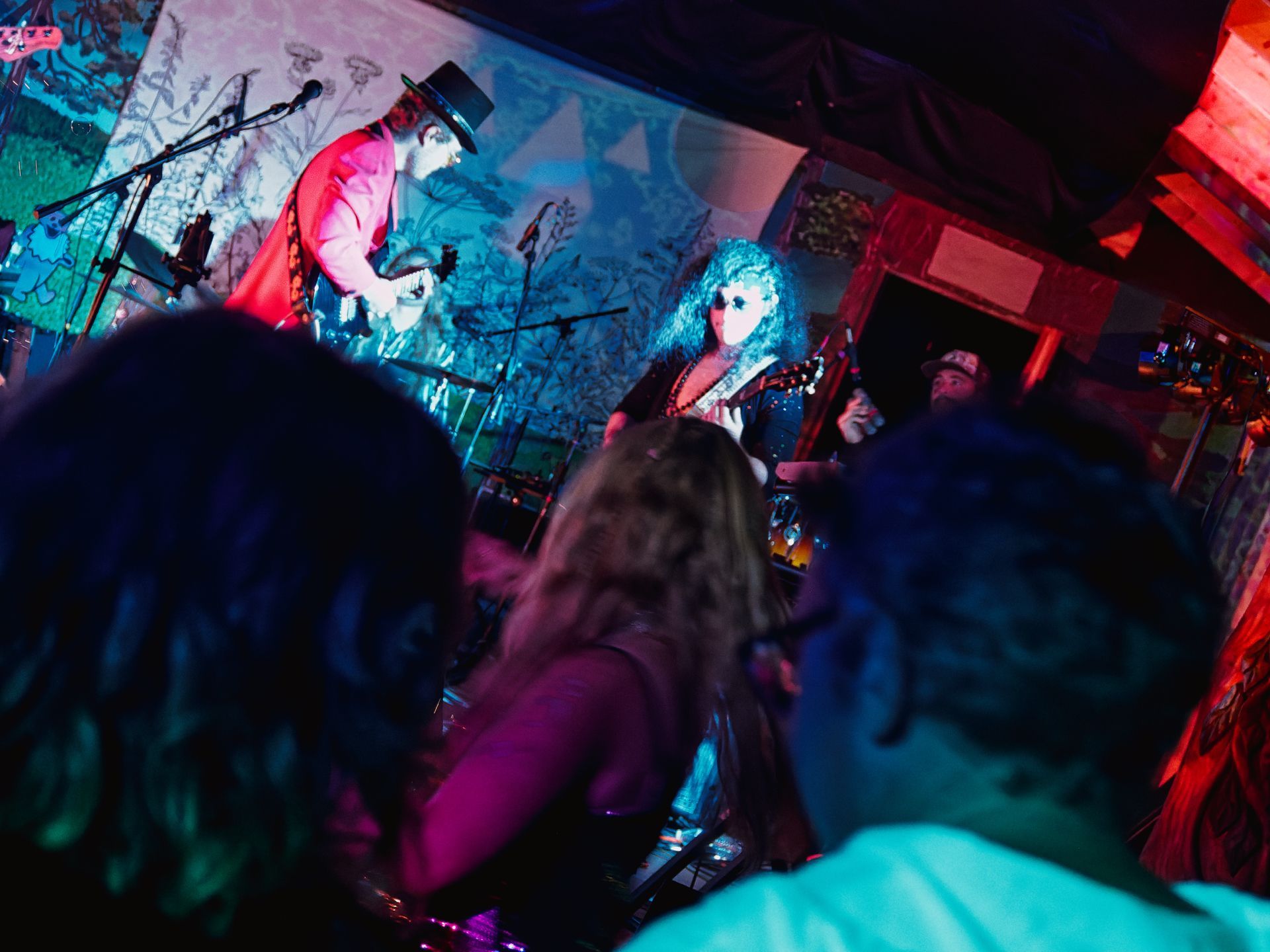 Two guitarists perform on a small, vibrantly lit stage — one in a red jacket and black top hat leaning into his guitar on the left, the other with voluminous curly hair bathed in teal light at center — while a crowd of blurred heads fills the foreground, shot from behind the audience.