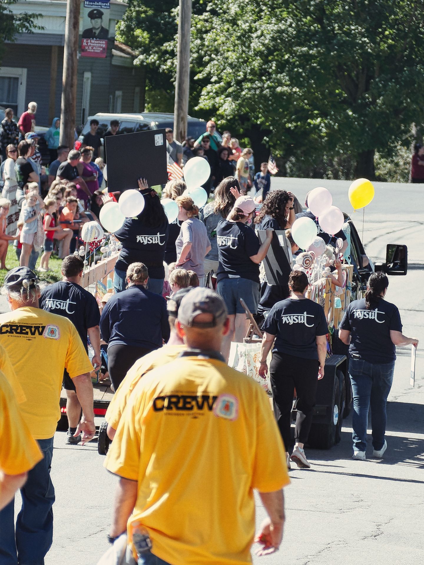 Vertical shot of a street parade in progress, with a group of people in navy "NYSUT" shirts walking alongside a decorated float carrying white, teal, and yellow balloons. In the foreground, two people in bright yellow "CREW / Stronger Together" shirts walk toward the procession, while a crowd of spectators lines the sidewalk to the left beneath full green trees.