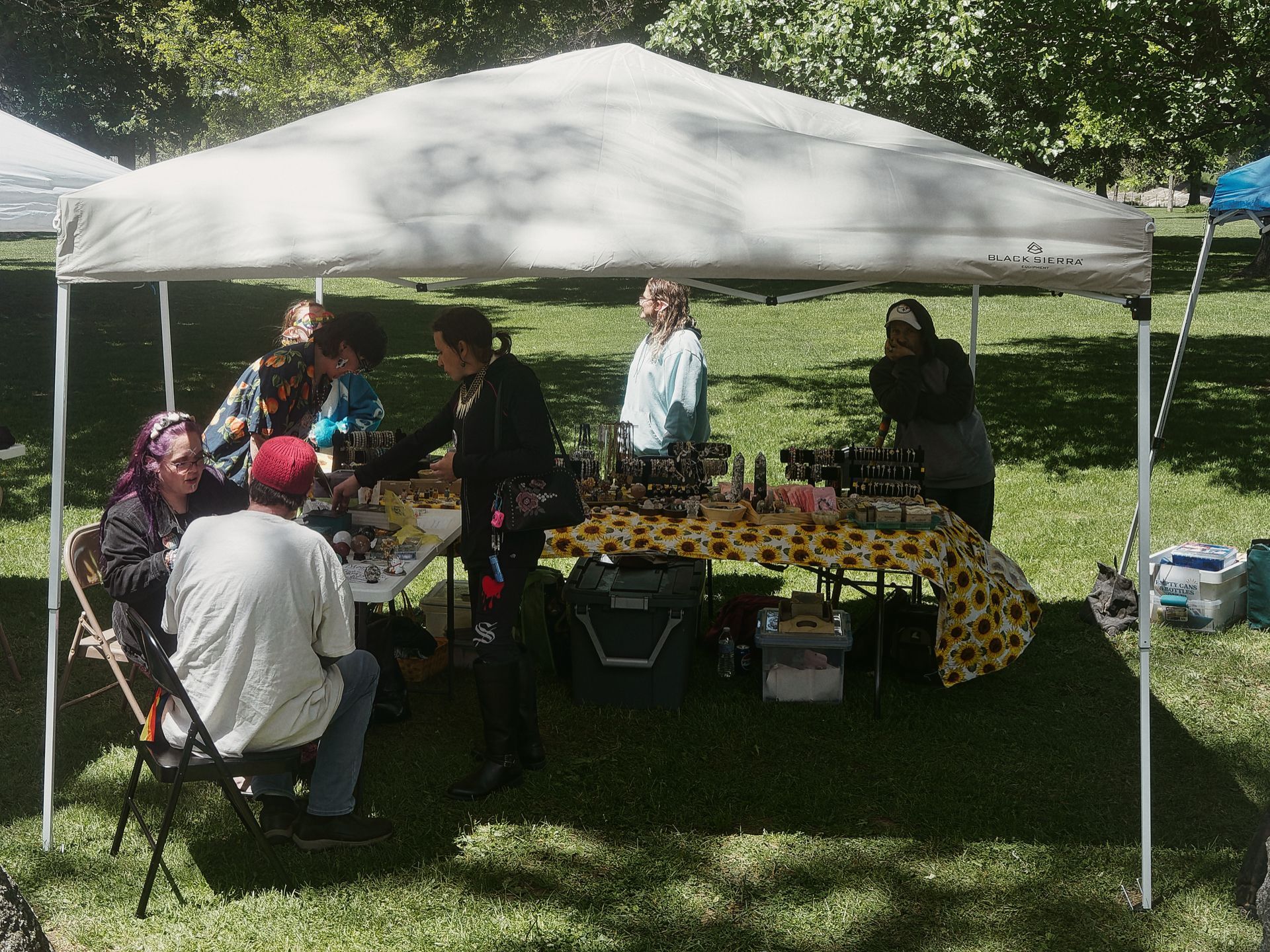 Outdoor market booth under a white Black Sierra pop-up canopy on a sunny grass lawn, with a vendor table covered in a sunflower-print yellow cloth and crowded with small jewelry and crystal items. Several people browse or sit around the table — including a person with purple hair seated at left and someone in a red beanie with their back to the camera — while leafy trees fill the background.