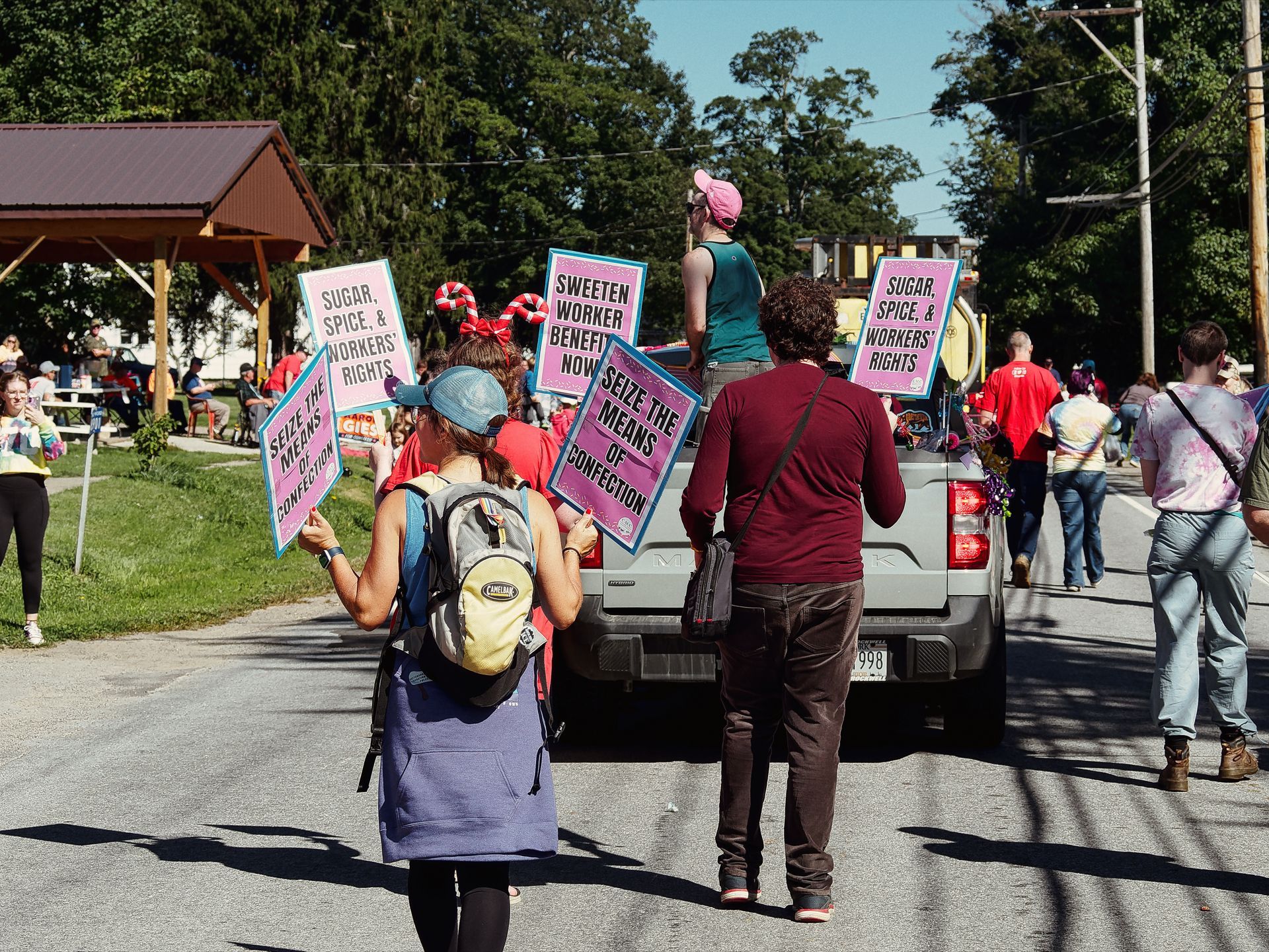 Parade participants walk behind a pickup truck down a tree-lined small-town road, carrying bright pink signs reading "SUGAR, SPICE, & WORKERS' RIGHTS," "SWEETEN WORKER BENEFITS NOW," and "SEIZE THE MEANS OF CONFECTION." A person in candy-cane headbands and a blue cap with a yellow backpack leads the foreground, while a participant perches atop the truck bed in a pink cap; onlookers and more marchers fill the street and grassy sideline to the right.