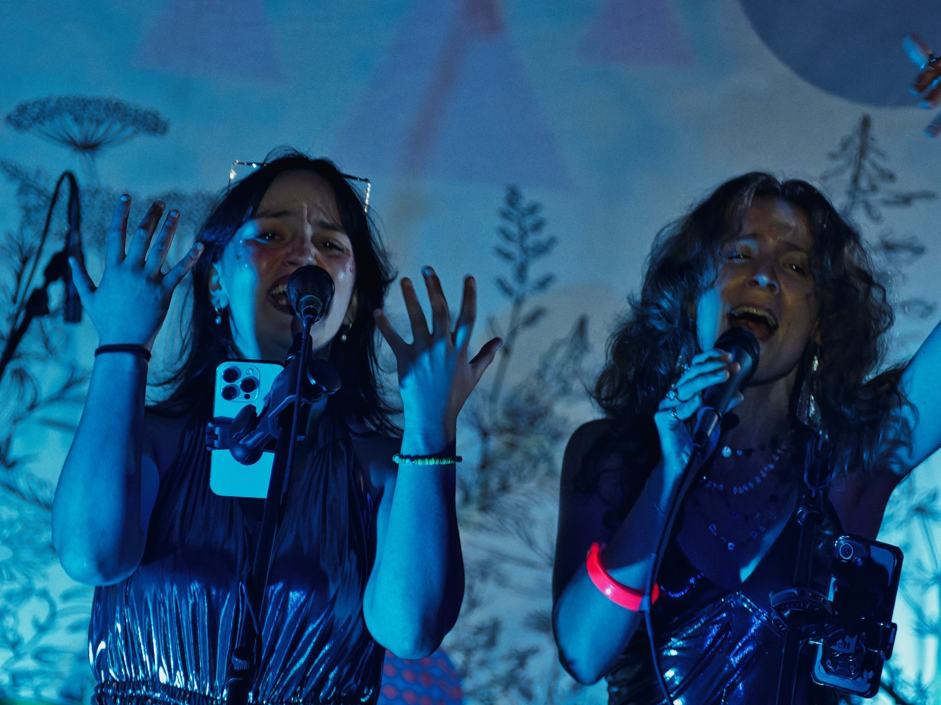 Two performers singing into microphones under deep blue stage lighting, both with hands raised and mouths open mid-song — the one on the left in a silver metallic top with a phone clipped to her outfit, the one on the right wearing a glowing red wristband and a GoPro-style camera at her hip. A botanical illustrated backdrop of silhouetted plants and trees fills the background.