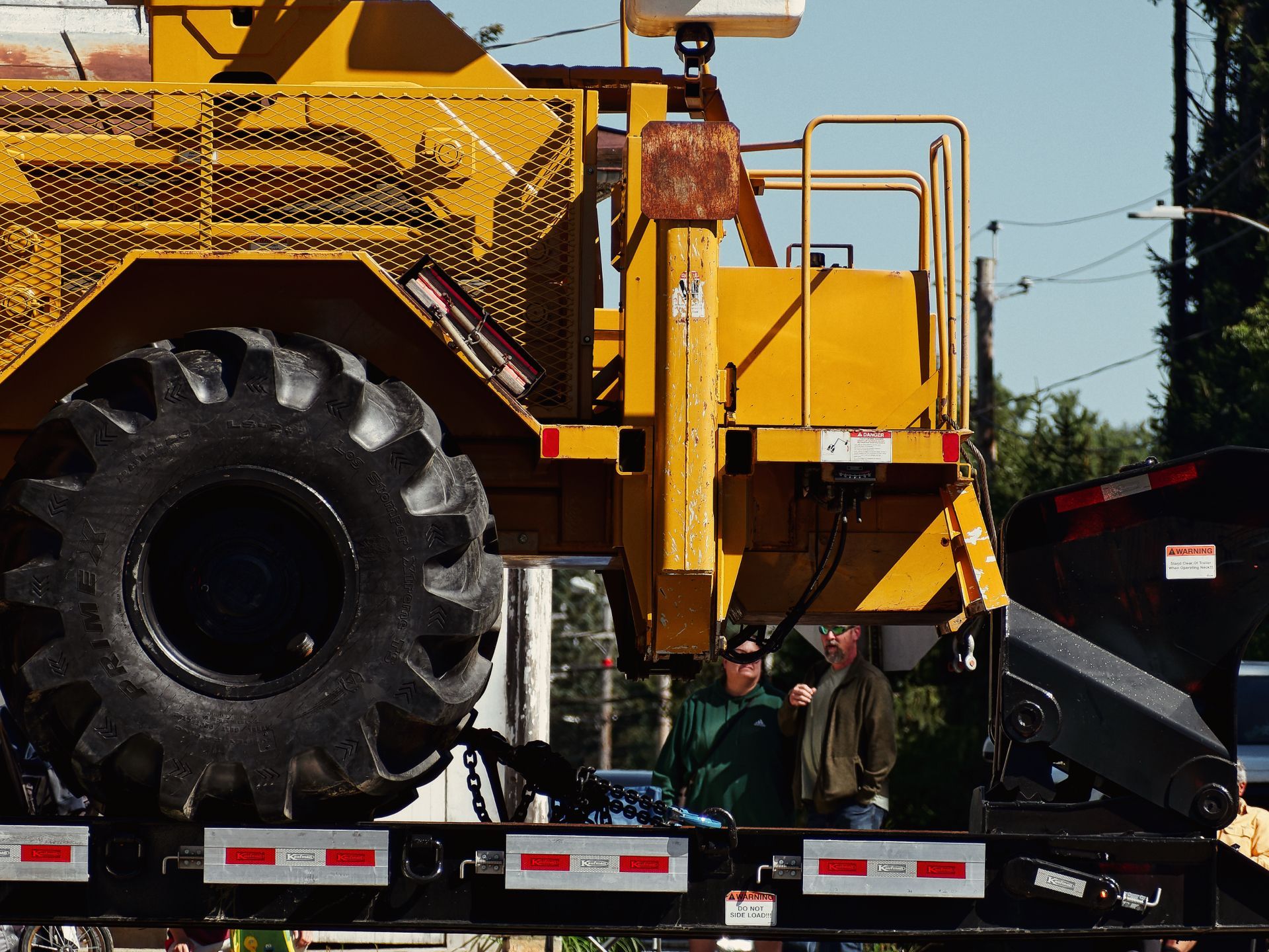 A massive yellow construction vehicle — its oversized knobby black tire dominating the left side of the frame — sits loaded on a flatbed trailer with red reflector strips along the edge. Two men stand partially obscured beneath the equipment in the midground, with trees, power lines, and a pale blue sky visible behind them to the right.