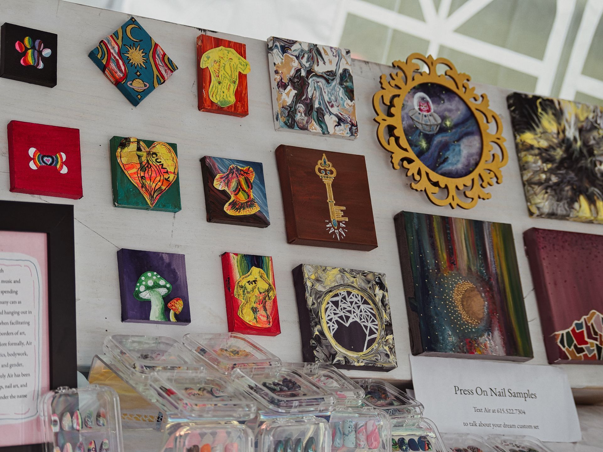 An art vendor's display wall covered in small colorful paintings — mushrooms, a glowing key on dark wood, a heart, a cosmic figure in an ornate gold frame, abstract pour paintings, and more — above a row of clear acrylic cases holding press-on nail samples. A small sign in the lower right reads "Press On Nail Samples / Text Air at 615.522.7304 / to talk about your dream custom set."