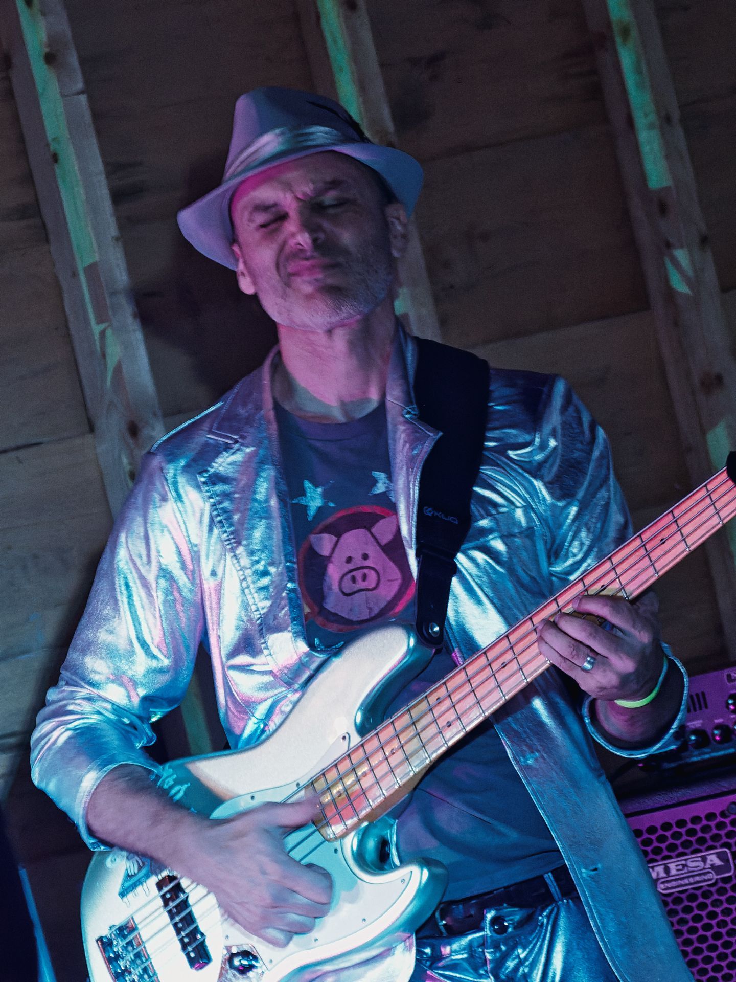 Vertical shot of a man in a silver metallic jacket and white fedora playing a pale green electric bass guitar, eyes closed mid-performance, lit in cool blue and pink stage lighting. His gray t-shirt features a cartoon pig face, and a Mesa Engineering amp glows purple in the background behind him against raw wood-plank walls.