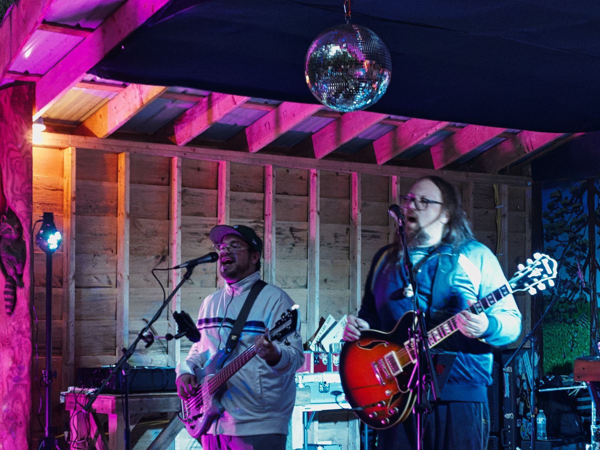 Two guitarists performing on a small wood-paneled stage bathed in pink and purple light, a mirror ball hanging from the exposed-beam ceiling above them — the one on the left wearing a backwards cap and singing into a mic, the one on the right with long hair and glasses playing a sunburst semi-hollow body guitar.