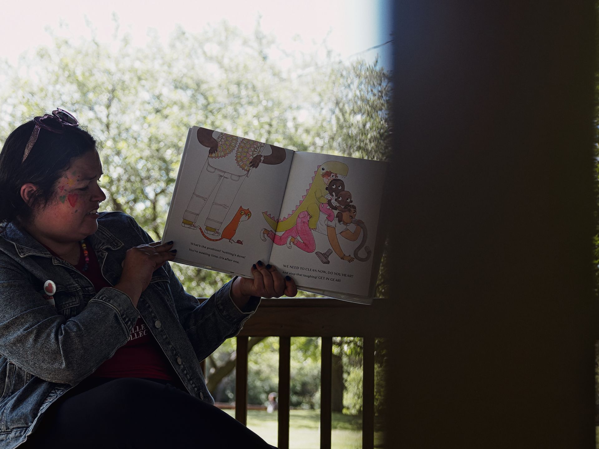 A woman in a denim jacket with face paint and sunglasses pushed up on her head reads an open children's picture book on a shaded porch, trees and a sunlit yard blurred softly behind her. The right page shows illustrated cartoon animals and a dinosaur-costumed figure, with text reading "WE NEED TO CLEAN NOW, DO YOU HEAR! And stop that laughing! GET IN GEAR!"