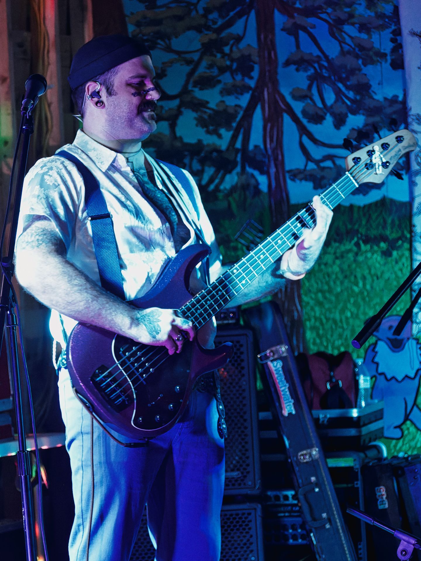 Vertical shot of a bassist in a flat-brim cap, suspenders, and a floral short-sleeve shirt, playing a dark five-string bass guitar under blue-purple stage lighting, facing slightly right with eyes downcast. Behind him, a vivid painted backdrop depicts tall redwood trees and rolling green hills, glowing under the UV wash.
