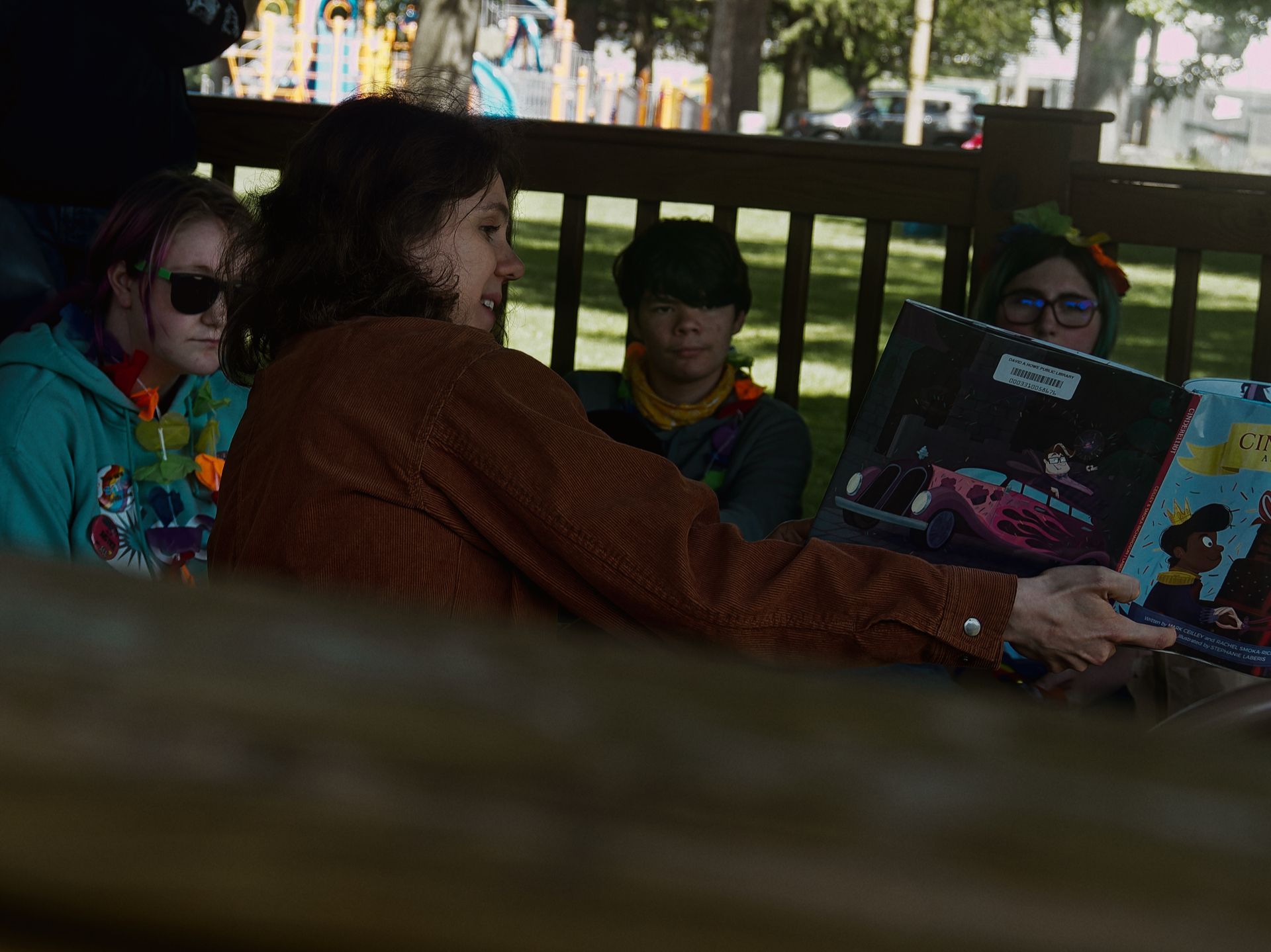 Person in a rust-brown corduroy jacket holding open a children's picture book — its spread showing an illustrated pink hot rod with flames — toward three attentive listeners seated on a wooden bench. The listeners wear colorful flower leis and accessories; the one on the left has pink-streaked hair and dark sunglasses. A sunlit playground and trees are visible through the bench railing in the background.
