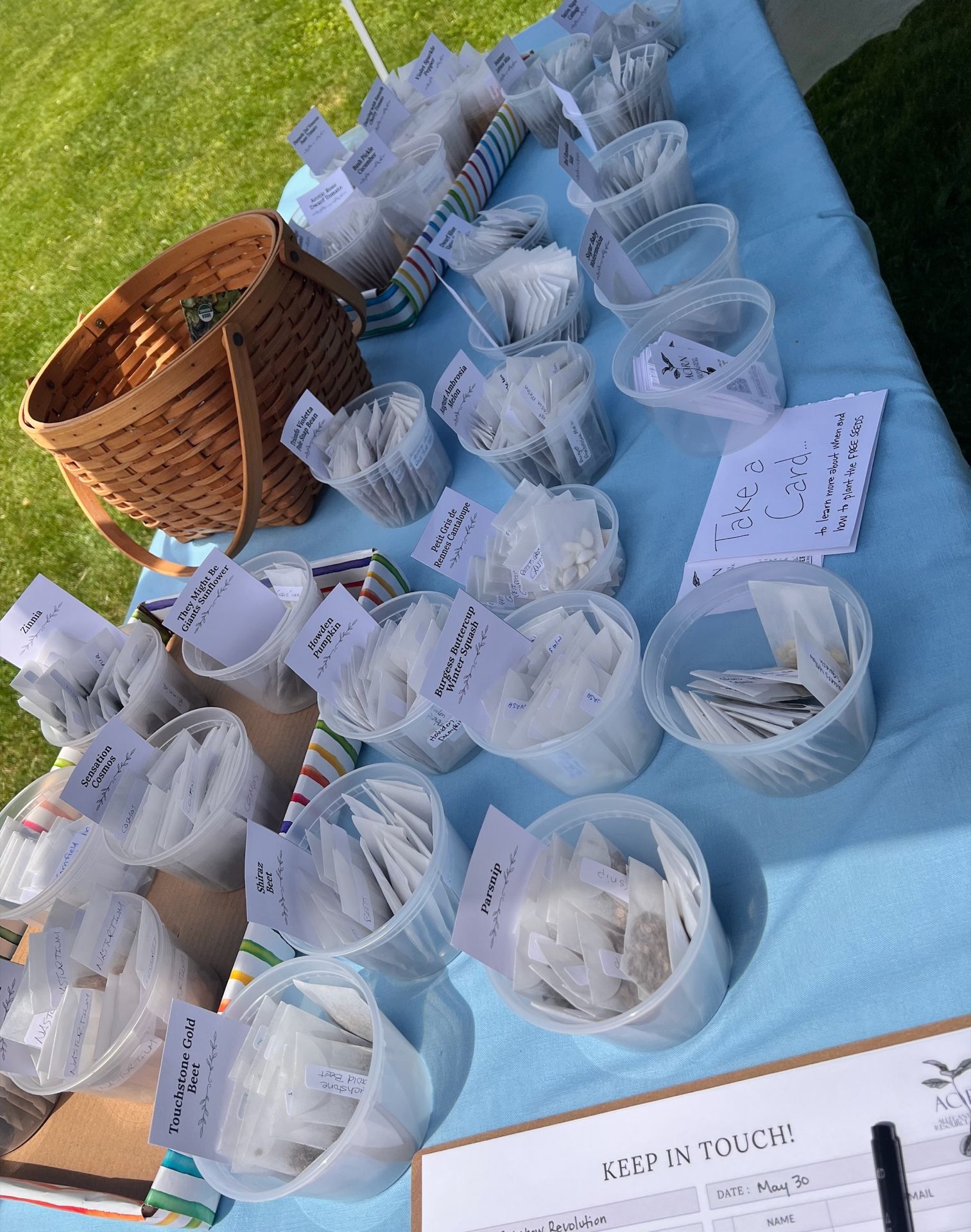 Overhead view of a light blue tablecloth covered in clear plastic cups filled with free seed packets, each labeled with handwritten variety names including "Parsnip," "Touchstone Gold Beet," "Shira Beet," "Sensation Cosmos," and "Zinnia," among others. A woven wicker basket sits to the left, a handwritten card reads "Take a Card… to learn more about when and how to plant the FREE seeds," and a sign-up sheet in the lower right corner reads "KEEP IN TOUCH!" with "DATE: May 30" filled in.