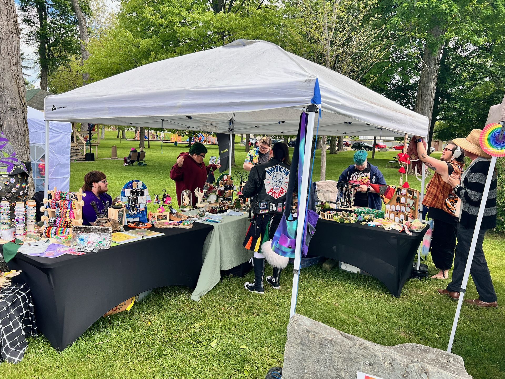 Outdoor craft fair booth under a white canopy tent on a grass lawn, tables draped in black and sage green cloth and crowded with colorful handmade jewelry, keychains, and small art pieces. Several vendors and shoppers browse the displays, including one person in a black jacket reading "American Psycho" and another on the right wearing a straw hat and headphones, with trees and a park green stretching into the background.