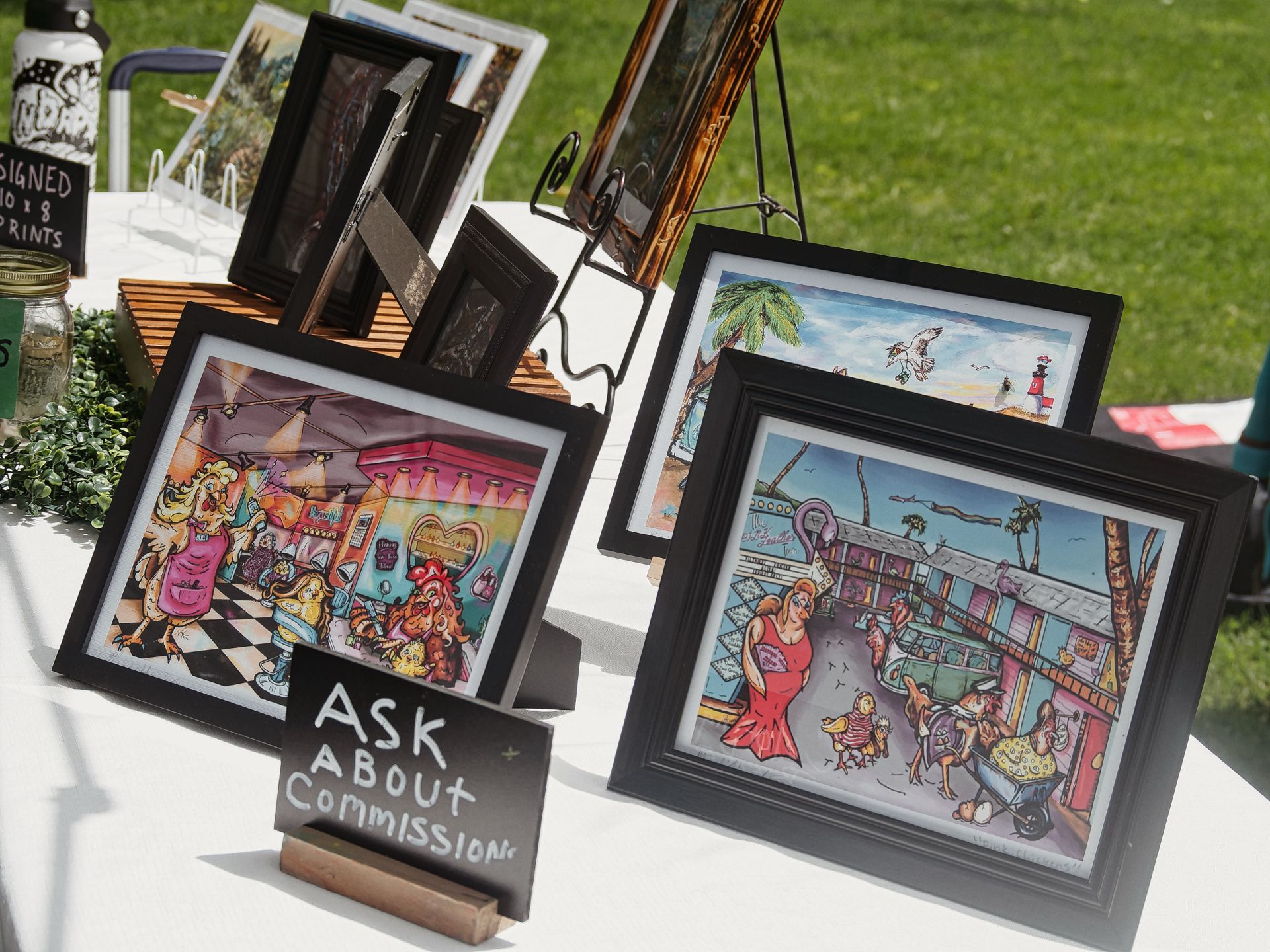 Outdoor art vendor table covered in white cloth, displaying several black-framed cartoon-style prints featuring whimsical characters — anthropomorphic chickens in a diner scene on the left, a vibrant beach boardwalk scene on the right. A small chalkboard sign in the foreground reads "ASK ABOUT Commissions." Additional framed prints and a sign reading "SIGNED 10 x 8 PRINTS" are visible at the left edge, with green grass blurred in the background.