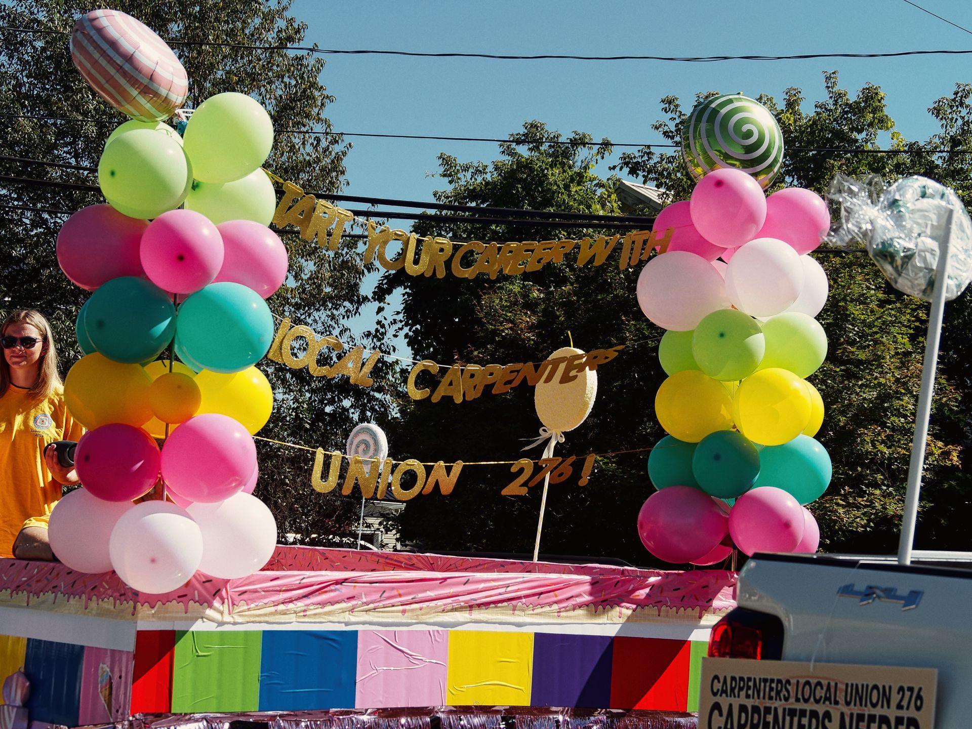 A parade float decorated with two arching columns of multicolored balloons — pink, teal, yellow, green, and white — topped with oversized candy-themed foil balloons. A gold glitter banner between the columns reads "START YOUR CAREER WITH LOCAL CARPENTERS UNION 276!" and a sign on the lower right reads "CARPENTERS LOCAL UNION 276 / CARPENTERS NEEDED." A woman in a yellow shirt and sunglasses stands to the left, holding a camera, with green trees and blue sky behind the float.
