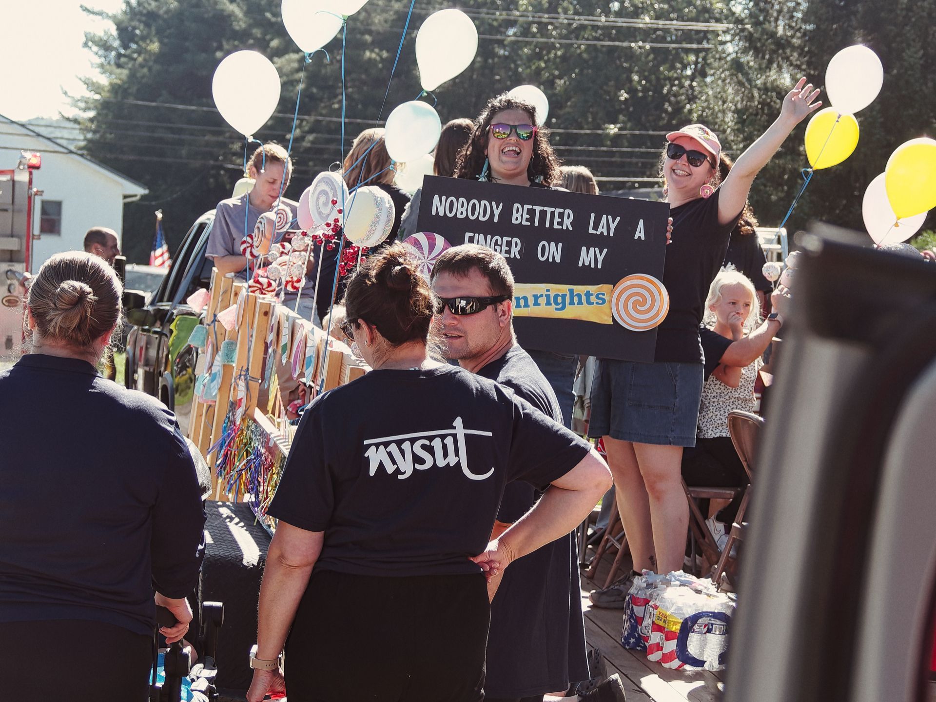 Outdoor parade scene with a candy-themed float decorated with oversized lollipop props and white and yellow balloons. A woman in sunglasses at center holds a black sign reading "NOBODY BETTER LAY A FINGER ON MY" followed by a partially obscured lollipop logo reading "nrights," riffing on the Butterfinger slogan; beside her, another woman waves with one arm raised. In the foreground, a person in a black t-shirt printed with "nysut" stands with their back to the camera.
