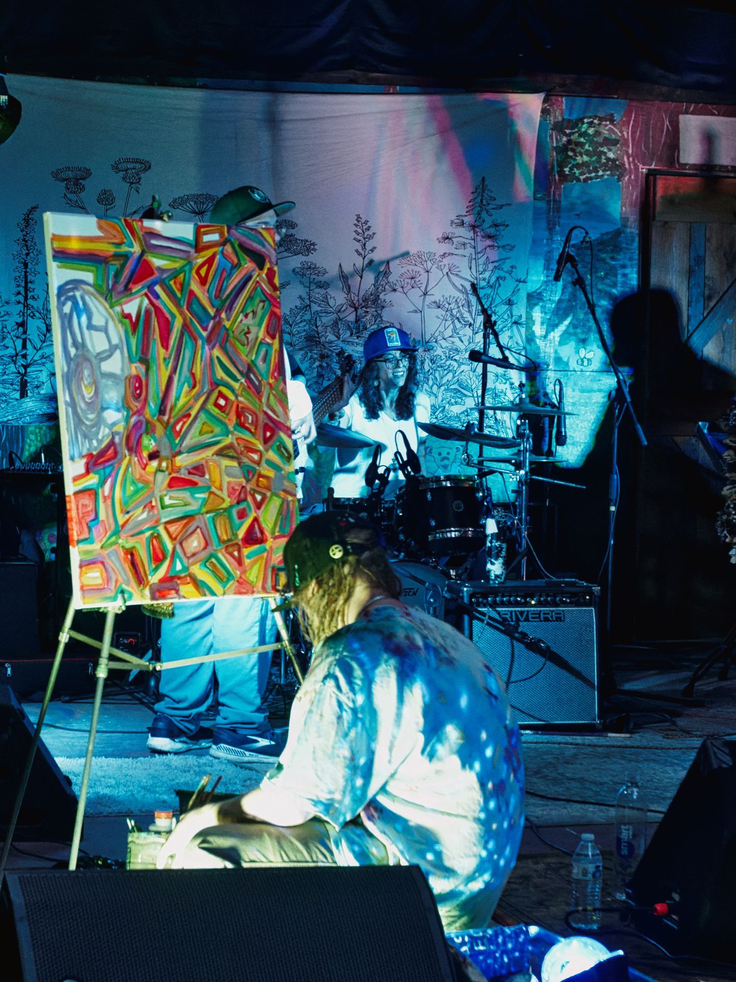 Vertical shot of a live performance bathed in deep blue stage lighting, with a large canvas on an easel in the foreground displaying a dense, colorful abstract painting of interlocking geometric shapes in red, green, yellow, and orange. A curly-haired artist crouches in front of it, bent low over their supplies, while a smiling drummer in a blue trucker hat plays a kit behind them. A silhouetted figure stands to the far right, and the backdrop features hand-drawn botanical illustrations with prismatic light projections washing across it.