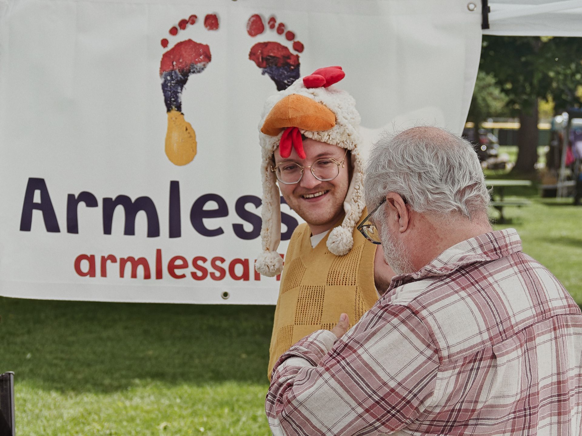 Two people mid-conversation at an outdoor event — a smiling young man in glasses wearing a plush chicken hat and a yellow knit vest, and an older gray-haired man in a red plaid shirt with his back to the camera. Behind them, a large white banner reads "Armless" in navy and "armlessar..." in red, partially obscured, with two colorful painted footprints above the text.