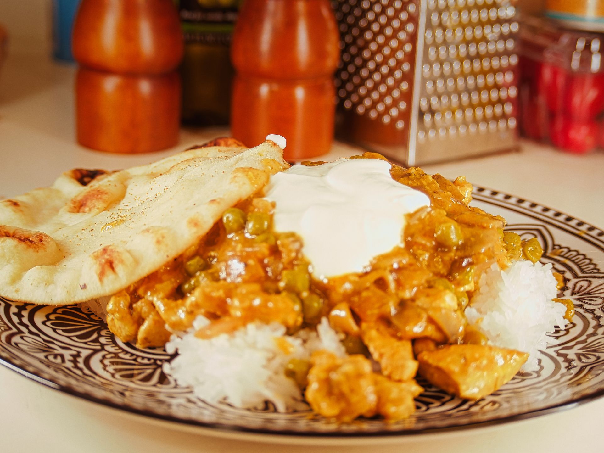 Golden-orange chicken and pea curry topped with a dollop of white yogurt, served alongside steamed white rice and a charred, blistered naan on a decorative brown-and-white patterned plate. Red salt and pepper shakers, a bottle of oil, a box grater, and a spice jar sit blurred in the shallow depth of the background.