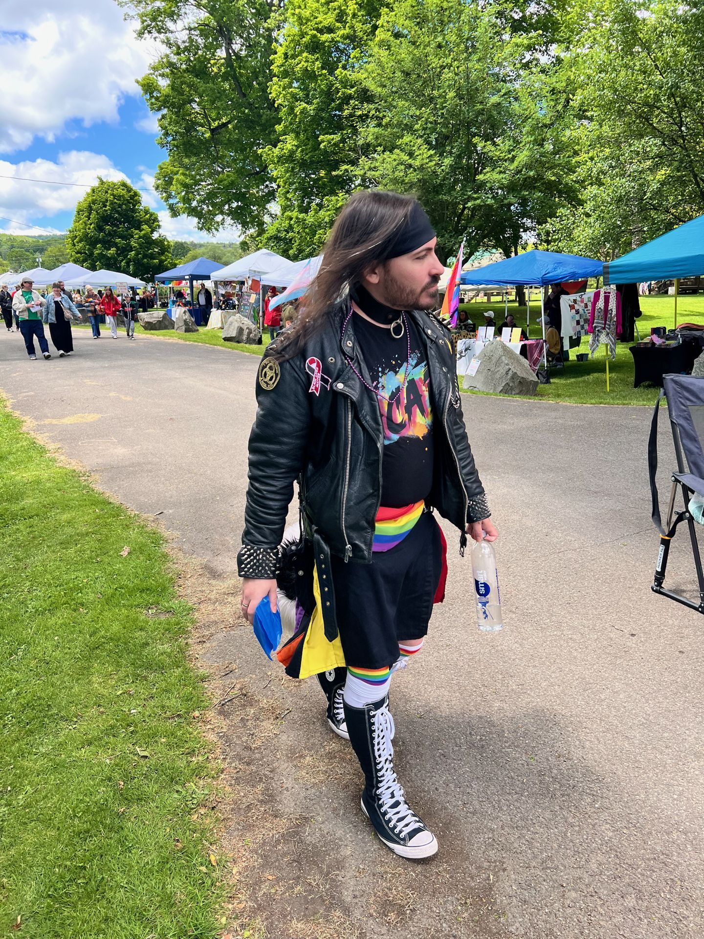Person with long dark hair and a black headband walking along a paved path at an outdoor Pride event, wearing a black leather jacket with pins, a rainbow-striped skirt over black shorts, rainbow knee-high socks, and tall black Converse high-tops, carrying a water bottle in one hand and a small rainbow flag in the other. Vendor tents in blue and teal line the right background under full green trees and a partly cloudy sky.