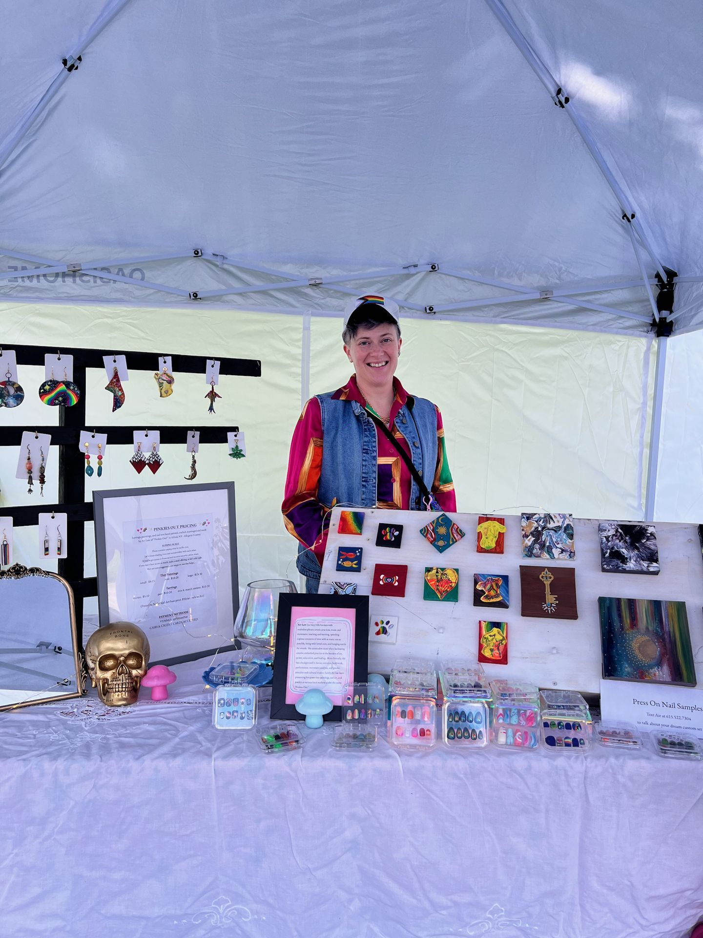 Vertical shot of a smiling vendor standing behind a craft fair table under a white canopy tent, wearing a colorful striped shirt, denim vest, and a rainbow-banded cap. The table is densely arranged with press-on nail sample sets in clear plastic cases, a gold skull figurine, small mushroom figurines, and framed pricing and artist info signs — one labeled "Press On Nail Samples" — while behind her, a black display board holds colorful dangling earrings and a white board displays small painted canvases featuring suns, moons, paw prints, keys, and abstract designs.