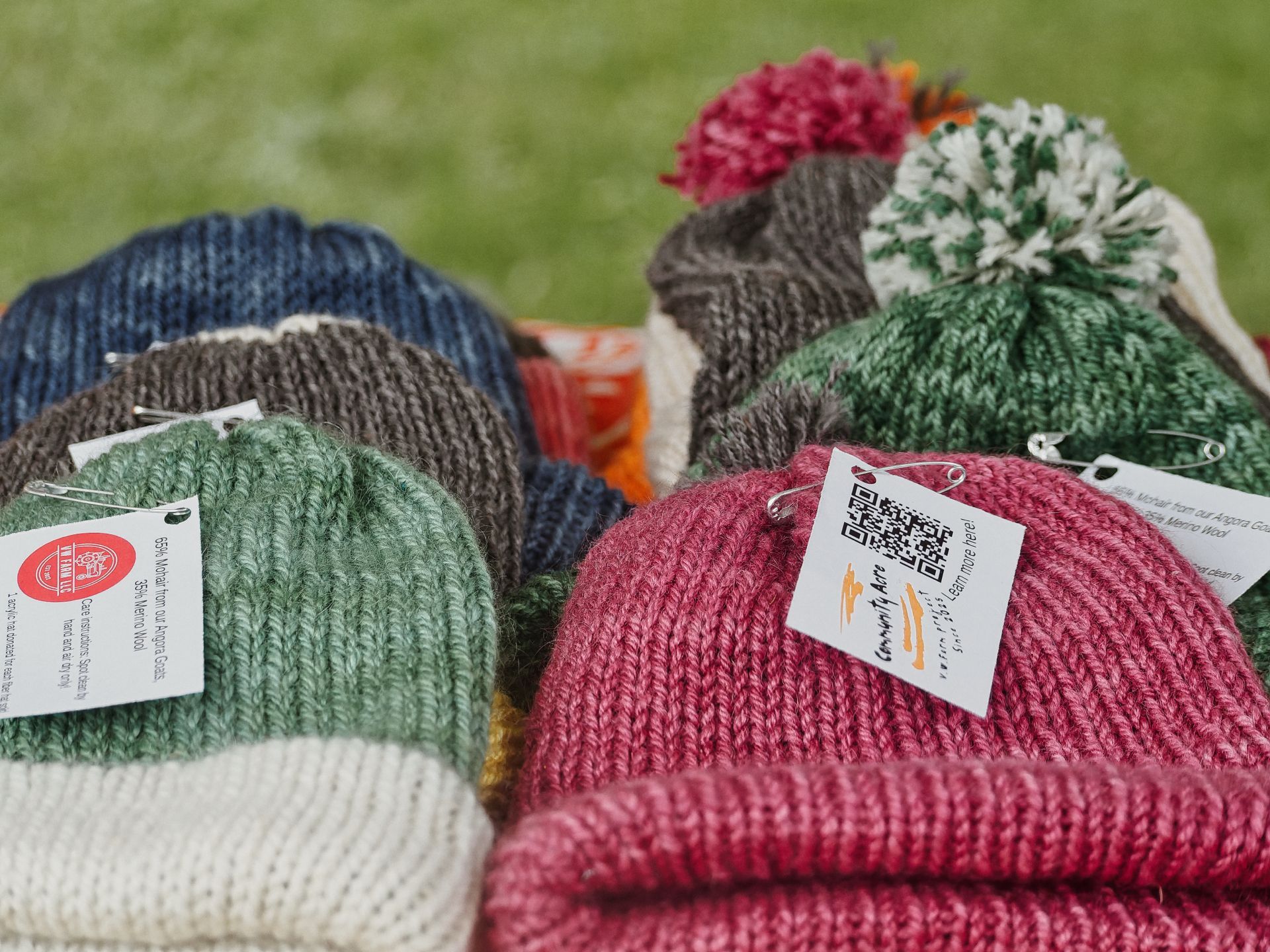Several hand-knit wool beanies in green, blue, grey, cream, and deep pink arranged in two loose rows on a grassy surface, each pinned with a small tag reading "Community Acre" with a QR code, and "65% Merino Wool" visible on the leftmost tag — two pom-pom hats in grey-pink and green-white rise above the others in the background.