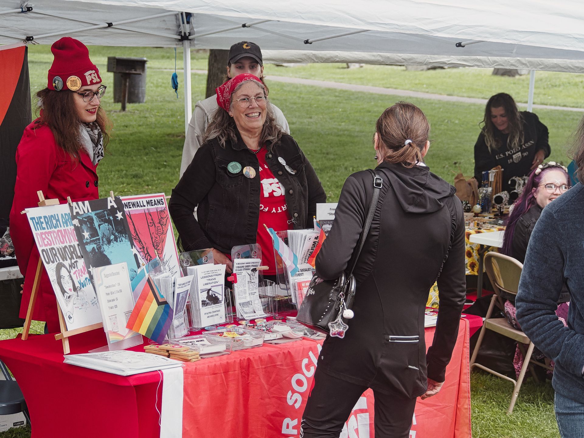 An outdoor event booth under a white canopy, draped in red tablecloths reading "SOCIALIST" along the front, covered in pamphlets, buttons, and a small rainbow Pride flag. A smiling woman in a red bandana, black vest with pins, and a red "PSL" shirt chats with a visitor in all black, while a younger woman in a red coat and red "PSL" beanie stands to the left beside a sign reading "THE RICH ARE DESTROYING OUR WORLD" and another reading "STONEWALL MEANS." A grassy park stretches into the background, where two other attendees are visible at a neighboring table.