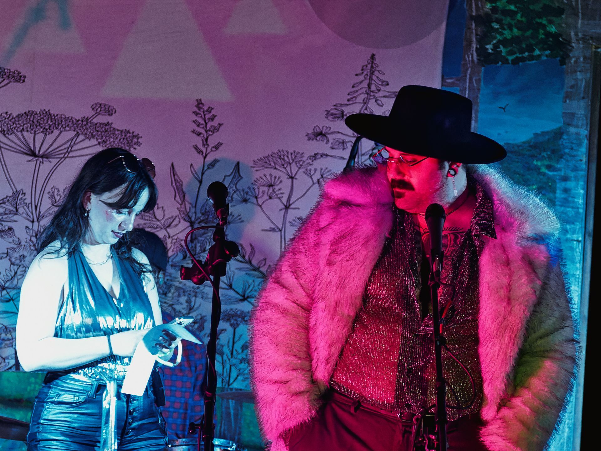 Two performers stand at microphone stands against a floral illustrated backdrop bathed in pink and blue stage lighting — on the left, a woman in a silver halter top and metallic jeans glances down at her phone, and on the right, a person in a wide-brimmed black hat, red glasses, a pink fur coat, and a glittery burgundy top leans slightly forward toward the mic.