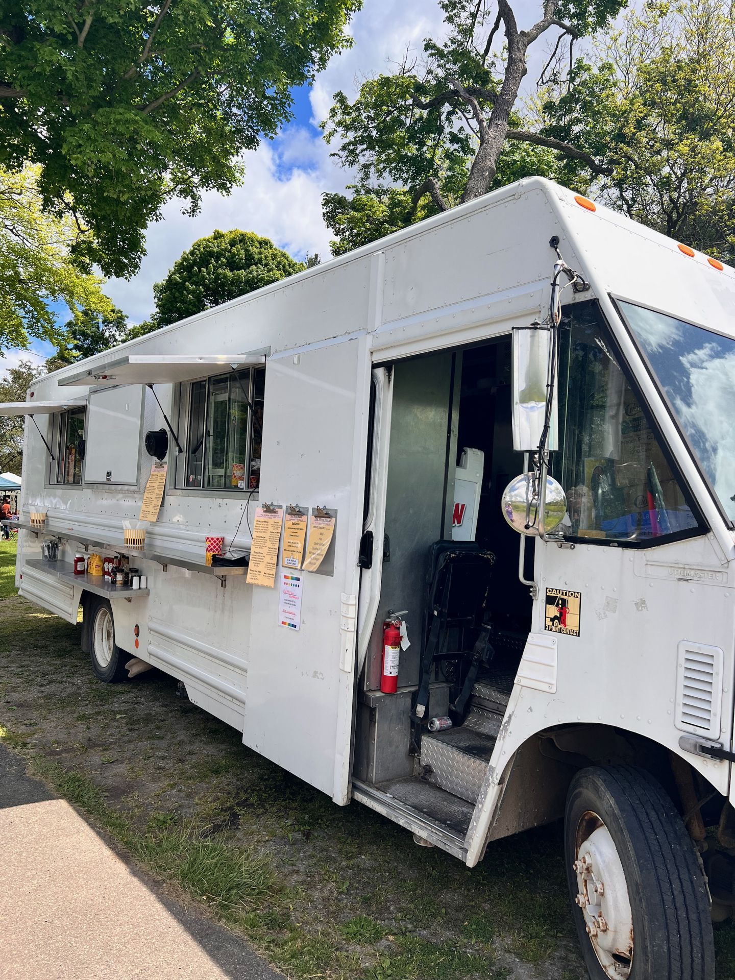 White food truck parked on a grassy lot beneath full green trees, its service window open on the left with a small counter, condiments, and several menu clipboards pinned to the side panel. The entry door on the right stands open, revealing interior steps and a red fire extinguisher mounted near the base, with a "CAUTION" sticker visible on the front cab corner.