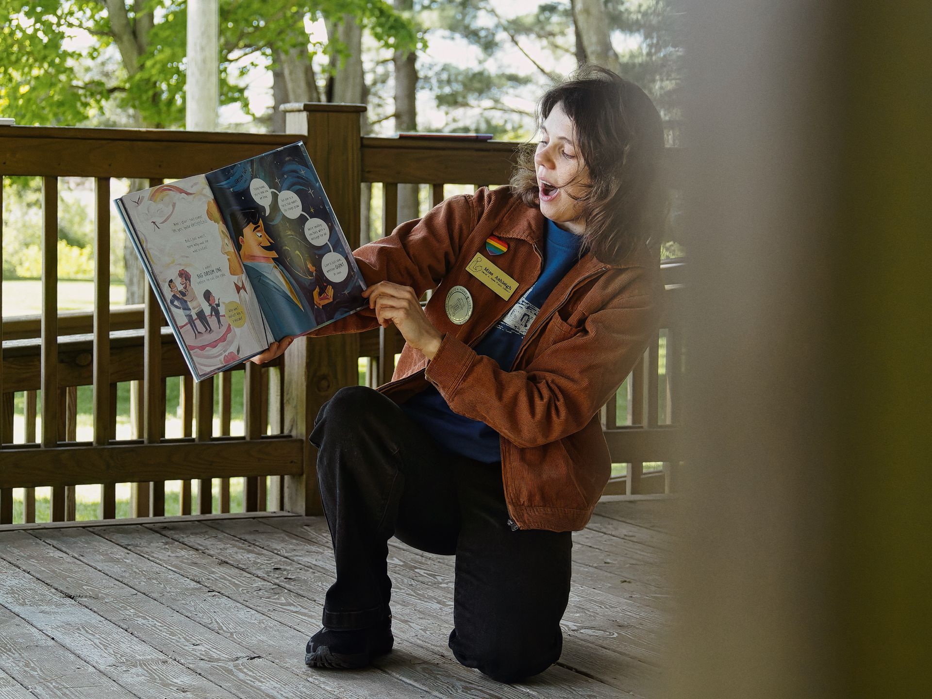 Woman in a rust-brown corduroy jacket — wearing a rainbow pin, a name badge, and a medallion — crouches on one knee on a wooden deck, mouth open mid-read, holding up an open illustrated children's book toward an unseen audience. The book's spread shows colorful cartoon figures with speech bubbles. Green trees and open lawn are visible through the railing behind her, slightly blurred in the background.